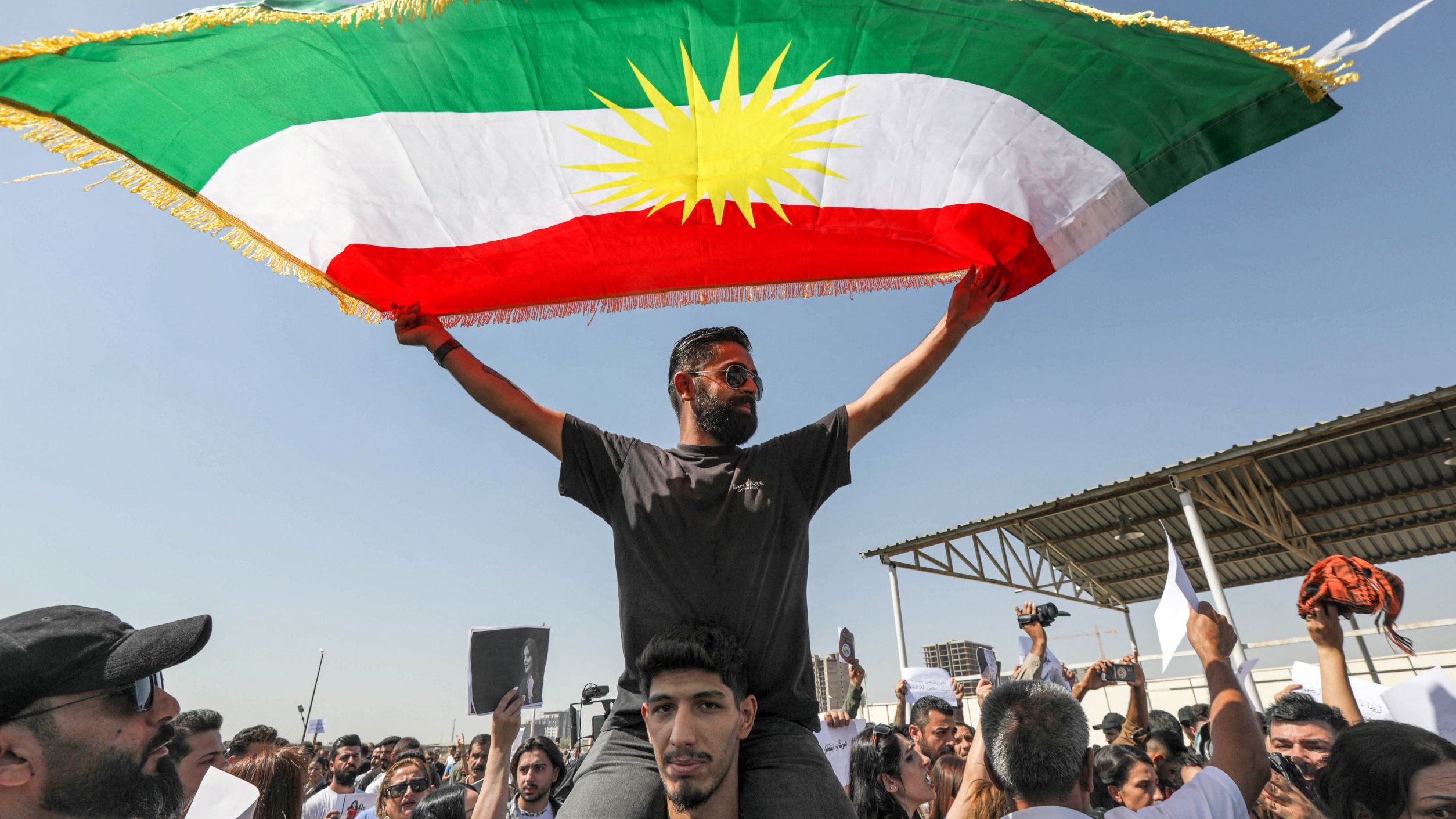 L'image montre un groupe de personnes lors d'une manifestation. Au premier plan, un homme se tient sur les épaules d'un autre, brandissant un grand drapeau kurde, qui est composé de bandes vertes, rouges et blanches avec un soleil jaune au centre. La foule autour d'eux paraît enthousiaste et engagée. Le ciel est clair, ce qui indique une journée ensoleillée. Des pancartes sont également visibles parmi les manifestants, suggérant qu'ils expriment des revendications ou des support pour une cause.