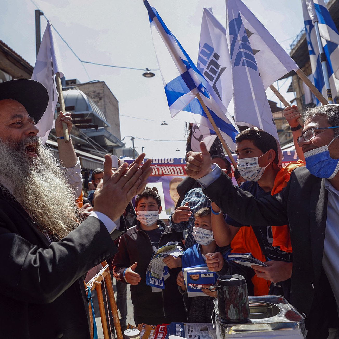 L'image montre une scène animée dans une rue, probablement lors d'un rassemblement ou d'une manifestation. Des personnes se tiennent debout, brandissant des drapeaux, apparemment en soutien à une cause. Au centre, un homme avec une longue barbe et un chapeau noir s'adresse à un autre homme, qui semble engagé dans un échange passionné. Les deux semblent entourés par une foule de jeunes gens, certains portant des masques de protection. L'environnement urbain et les bâtiments en arrière-plan ajoutent à l'atmosphère dynamique de l'événement.