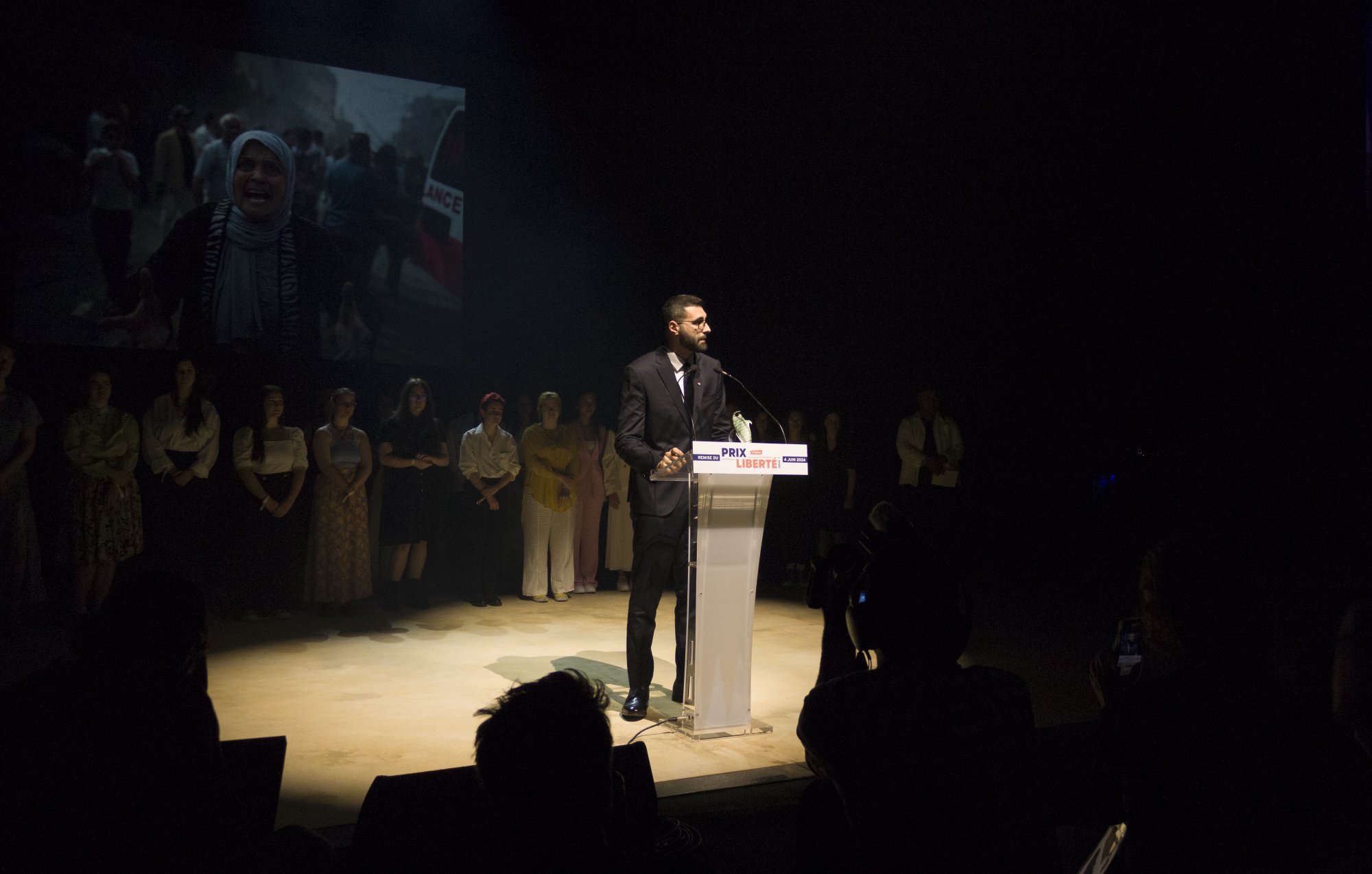 L'image montre un homme debout à un pupitre transparent, prenant la parole lors d'un événement. Il est habillé en costume et semble s'adresser à un public. En arrière-plan, on peut voir un écran projetant une vidéo ou une image, vraisemblablement liée à un sujet important, étant donné l'expression des personnes présentes. Au premier plan, on peut apercevoir des membres du public et des photographes, créant une atmosphère de concentration et d'engagement. L'éclairage met en évidence l'orateur, tandis que le reste de la scène est plus sombre.