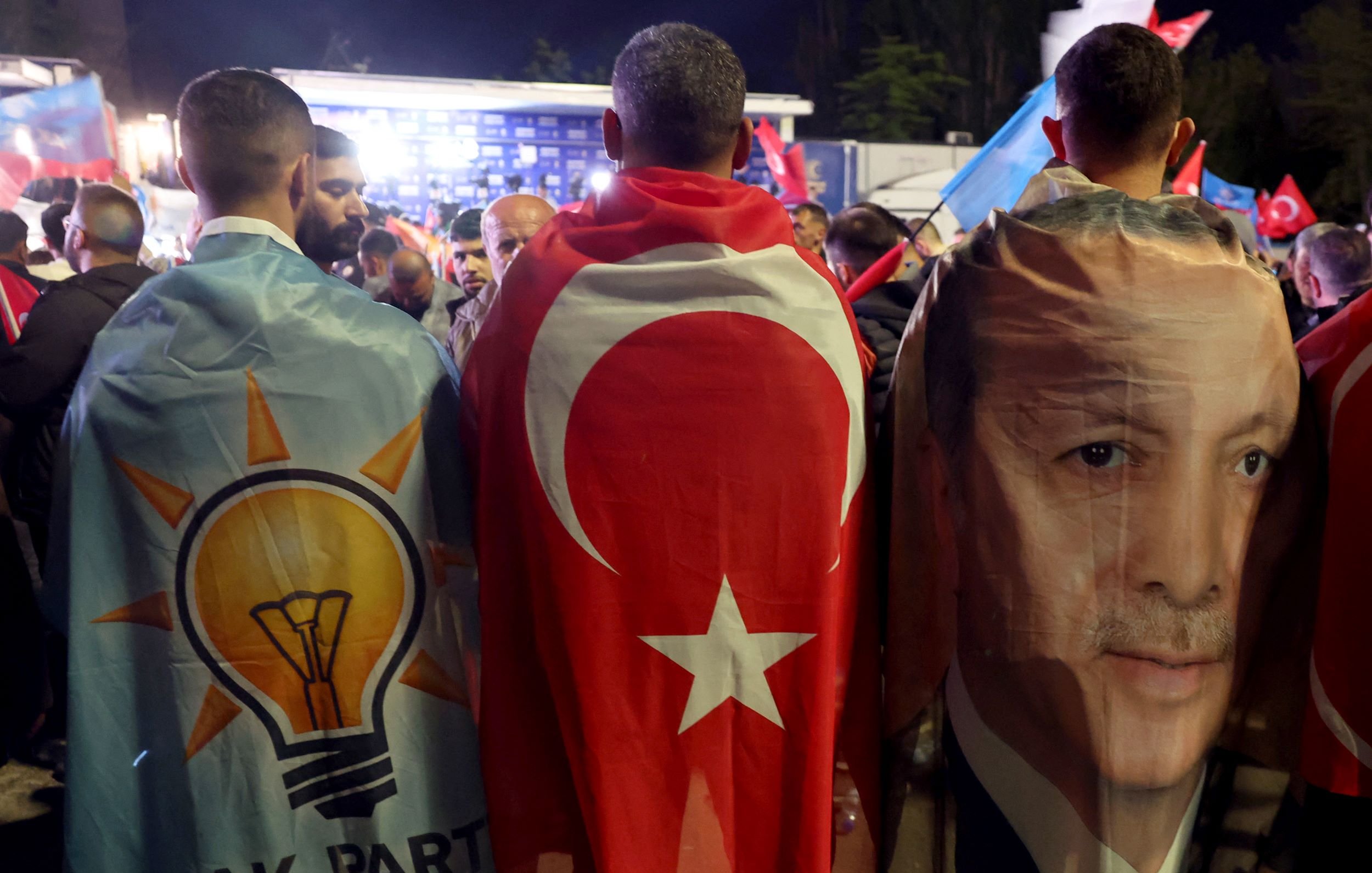 L'immagine mostra un gruppo di persone che indossano mantelli colorati. Due di loro hanno i mantelli decorati con il simbolo del partito AKP (Partito della Giustizia e dello Sviluppo) e la bandiera turca. Inoltre, uno dei mantelli presenta l'immagine di una figura pubblica, probabilmente un leader politico turco. Sullo sfondo si possono vedere altre bandiere e un'atmosfera di festa o celebrazione.