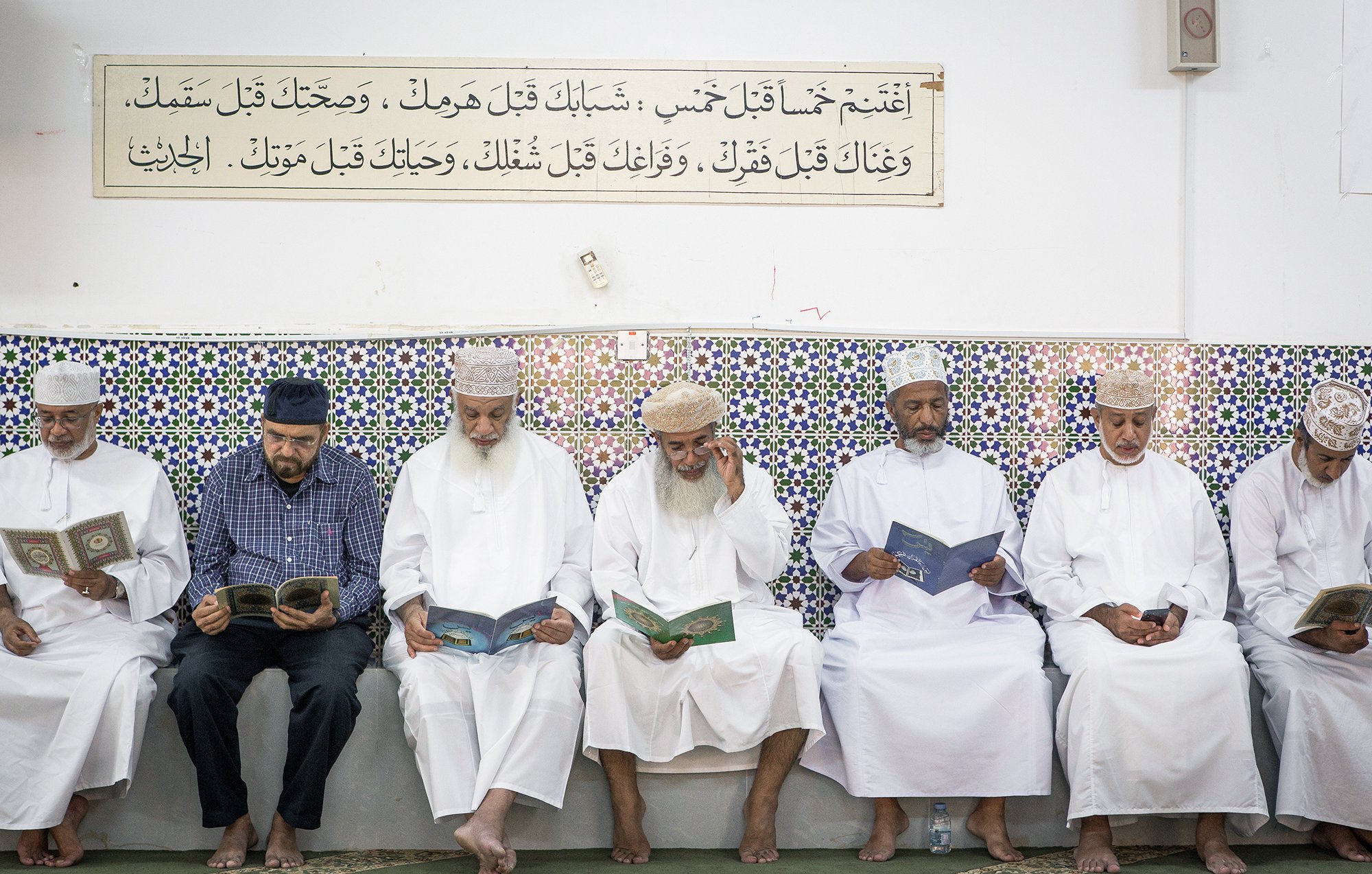 L'image montre un groupe d'hommes assis en ligne, lisant des livres, probablement des copies du Coran. Ils portent des vêtements traditionnels, tels que des dishdashas et des couvre-chefs. En arrière-plan, on peut voir un mur décoré de carrelage coloré, sur lequel est inscrite une citation ou un texte. L'atmosphère semble calme et sereine, suggérant un moment de prière ou de méditation.