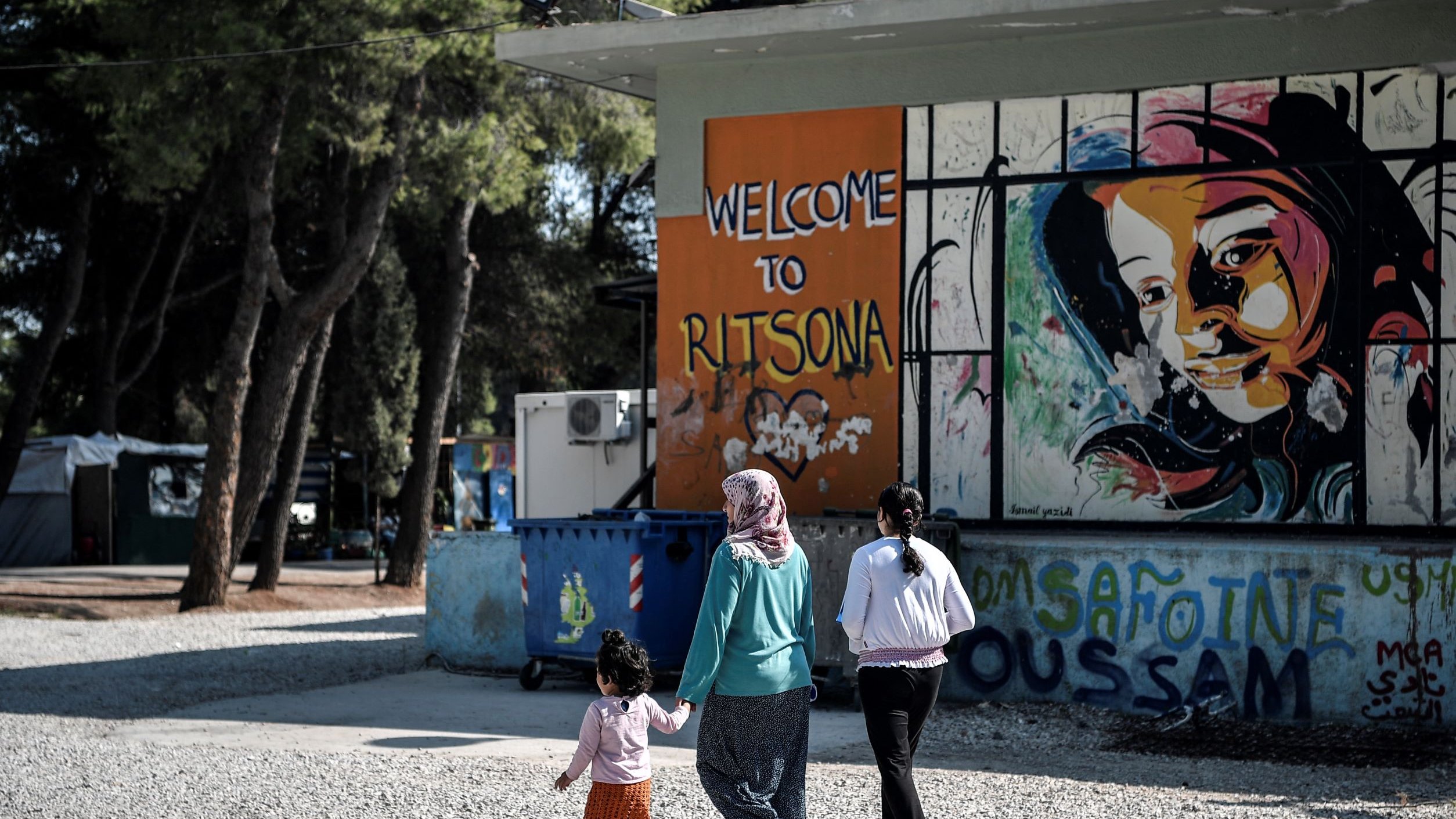 L'image montre un groupe de personnes marchant sur un chemin en gravier. À gauche, on peut voir un conteneur à ordures et à droite, un mur peint avec des graffitis colorés, dont le mot "Welcome to Ritsona" et le portrait d'une fille. Les personnes présentes semblent être une mère accompagnée de deux enfants, l'un d'eux tenant la main de la femme. L'environnement végétal suggère un cadre tranquille, malgré la situation.