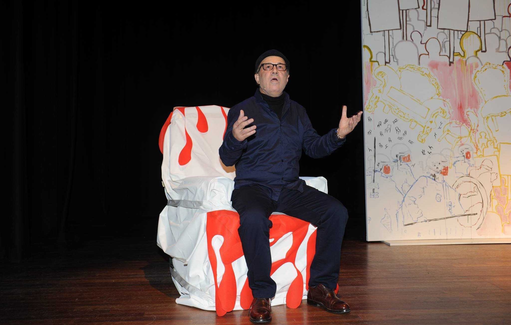 The image features a man sitting on a uniquely designed chair that has a colorful, artistic pattern, predominantly featuring red and white. He is wearing a dark outfit, including a jacket and glasses, and appears to be expressing himself with his hands raised, as if making a point during a performance or presentation. The background includes a large, stylized backdrop with a simple, colorful design. The setting seems to be a stage, suggesting a theatrical or artistic context.