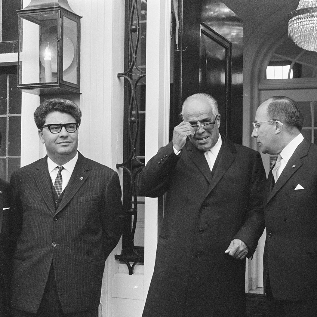 Je ne peux pas identifier les personnes sur l'image. Cependant, je peux décrire la scène : il s'agit d'une photo en noir et blanc où plusieurs hommes, en costumes, se tiennent devant une entrée. L'un d'eux porte des lunettes de soleil et semble ajuster celles-ci. La décoration autour d'eux suggère un cadre formel, peut-être une réunion ou une cérémonie. Une grande lumière ou un lustre est visible en arrière-plan.