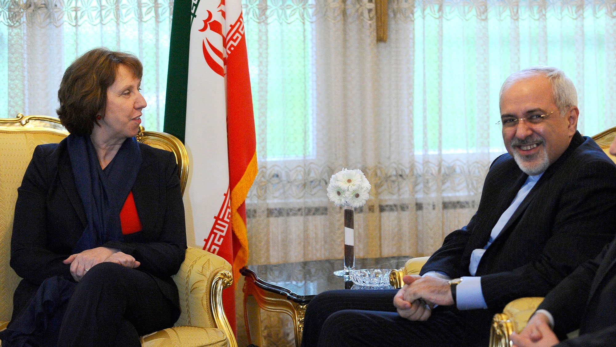 L'image montre une rencontre entre deux personnes assises sur des fauteuils dans un décor formel. À gauche, une femme avec des cheveux bruns, portant un blazer noir et un haut rouge, semble engagée dans la conversation. À droite, un homme souriant, vêtu d'un costume sombre, répond à la femme. En arrière-plan, on peut voir un drapeau iranien, ce qui suggère que la rencontre a lieu en Iran. La lumière naturelle entre par les fenêtres, créant une atmosphère accueillante.