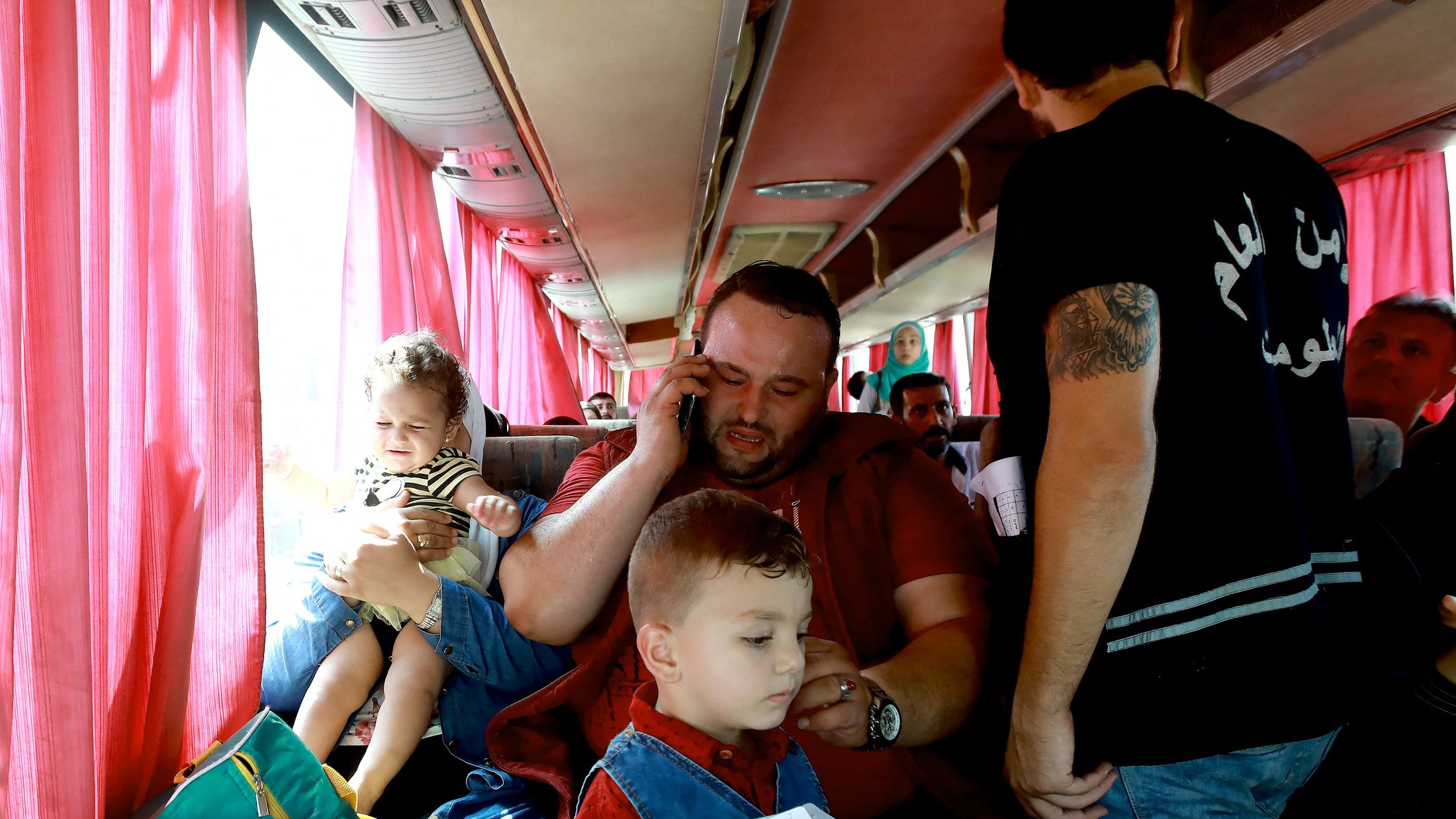 L'image montre un intérieur de bus avec plusieurs personnes. Un homme assis au centre tient un enfant dans ses bras et parle au téléphone. À ses pieds, un autre enfant semble jouer ou être attentif. D'autres passagers sont visibles autour, certains se tenant debout. Les sièges sont occupés, et des rideaux rouges laissent passer une lumière douce à travers les fenêtres. L'atmosphère semble chaleureuse et animée.