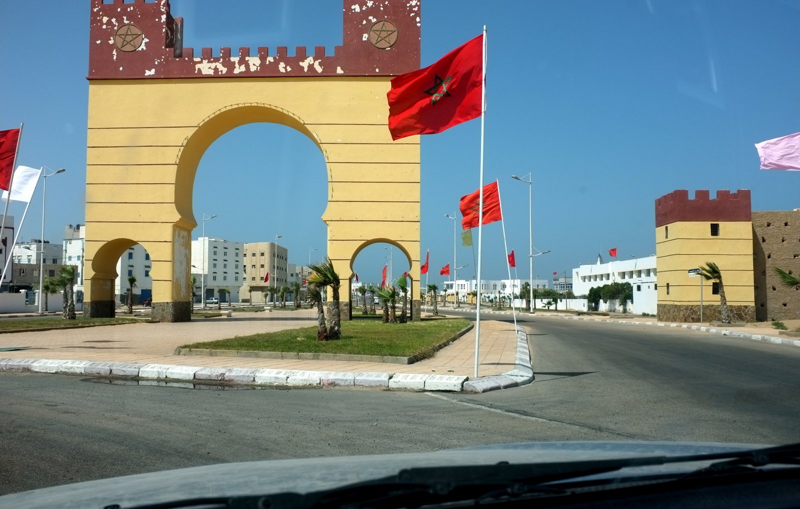 L'image montre un grand arc de triomphe peint en jaune avec des éléments rouges en haut, au centre d'une route. Drapeaux marocains flottent de chaque côté de l'arc, ajoutant une touche nationale à la scène. Des bâtiments modernes peuvent être aperçus au loin, tandis que la route est bordée de palmiers et d'une pelouse bien entretenue. Le ciel est clair et bleu, ce qui donne une ambiance ensoleillée et agréable à l'ensemble.