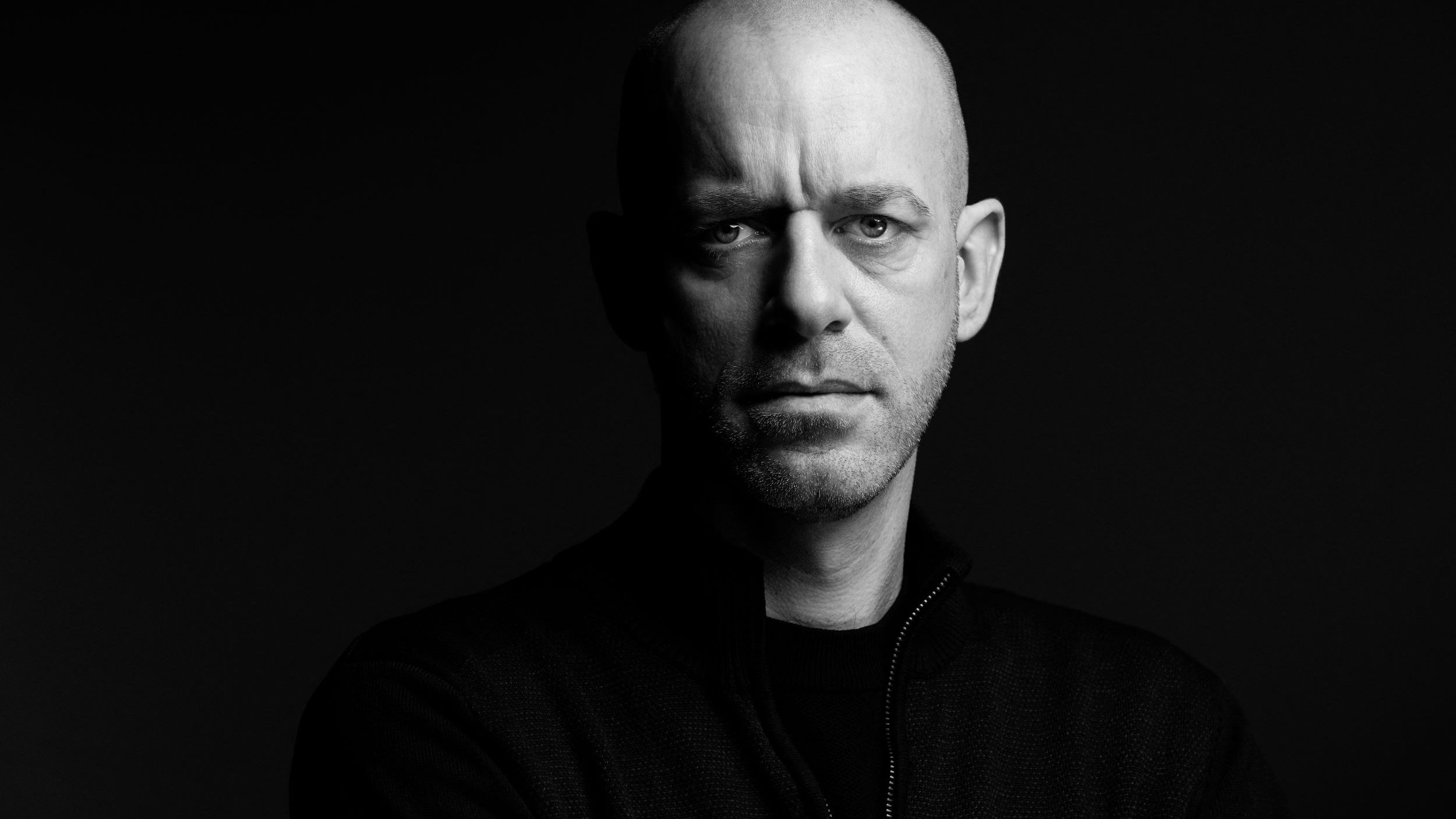 الصورة تُظهر رجلًا ذو رأس أصلع، يقف في ضوء خافت مما يبرز ملامح وجهه بوضوح. عيونه تحمل تعبيرًا جادًا وقويًا، بينما يحدق بشكل مباشر نحو الكاميرا. يرتدي سترة داكنة، ويبدو أن الخلفية سوداء، مما يضيف إلى جمالية الصورة ويعطي إحساسًا بالدراماتيكية.