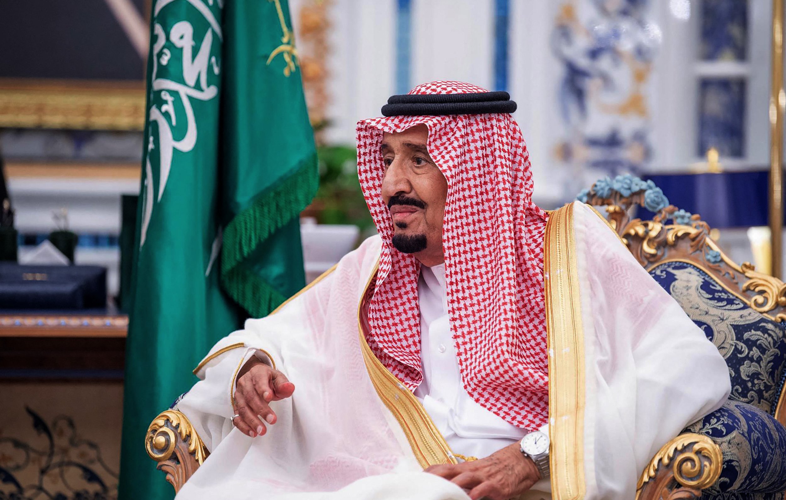 The image shows a man seated in a lavish decor room, wearing traditional attire that includes a white thobe and a red and white checked keffiyeh held in place by an agal. He has a thoughtful expression and is gesturing with his hand. Behind him, there is a green flag, likely representing Saudi Arabia, and opulent furnishings in the background.