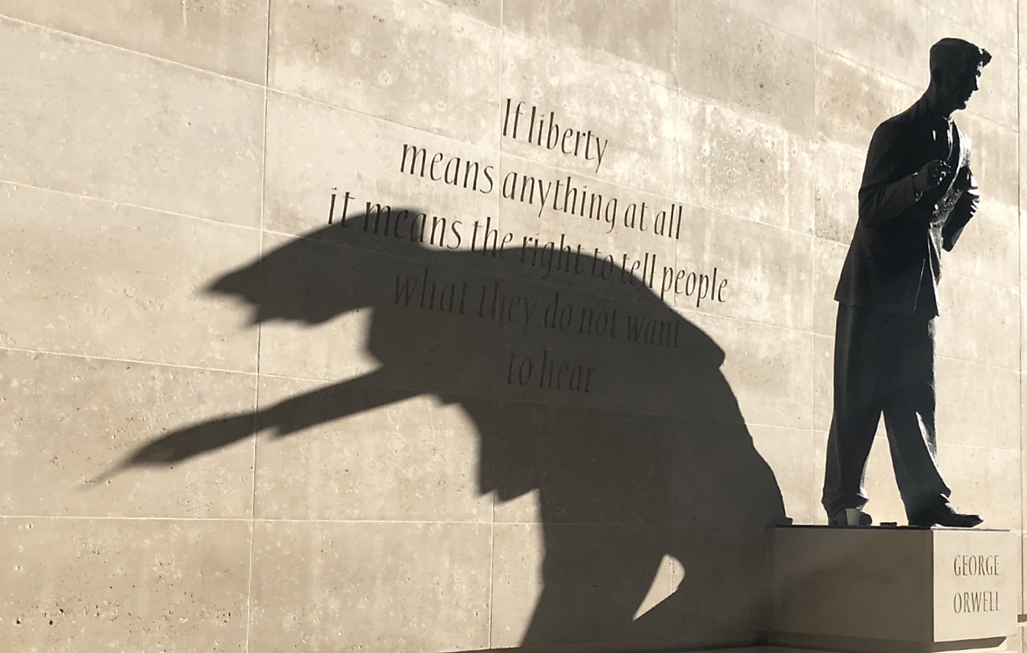 L'image montre une sculpture d'une silhouette d'un homme, probablement un écrivain, sur un socle avec le nom "GEORGE ORWELL". À côté de la statue, il y a une citation en anglais sur le thème de la liberté. La lumière crée une ombre qui semble projeter une forme dynamique, ajoutant une dimension artistique à la scène. Le mélange de texte et de sculpture évoque des réflexions sur la liberté d'expression.