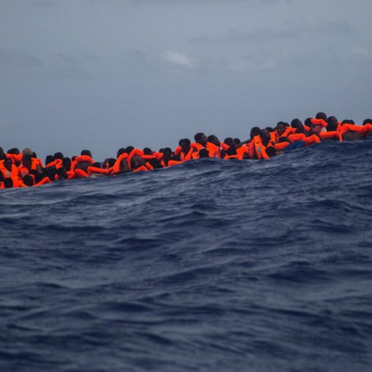 L'image montre un groupe de personnes en mer, portant des gilets de sauvetage orange. Elles semblent rassemblées sur une petite embarcation, luttant contre des vagues dans une étendue d'eau bleue profonde. Le ciel est nuageux, ce qui accentue la gravité de la situation maritime. Cette scène évoque des thèmes de migration, de survie et de danger en mer.