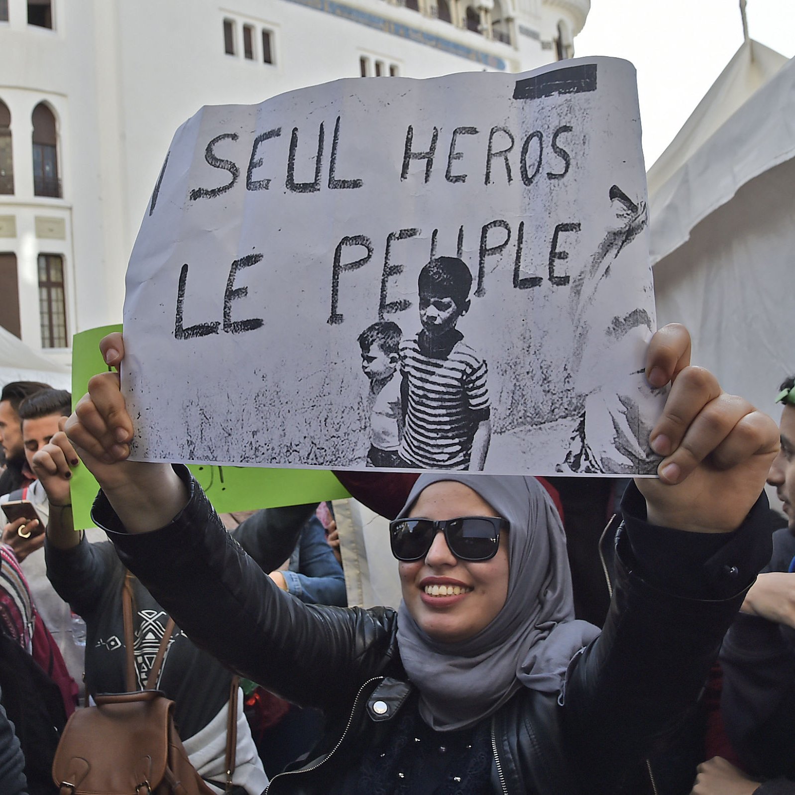 L'image montre une manifestation avec des personnes rassemblées. Au premier plan, une femme portant un hijab et des lunettes de soleil tient une pancarte qui affiche le slogan « SEUL HÉROS LE PEUPLE ? ». L'ambiance semble être engagée, avec d'autres manifestants en arrière-plan et des banderoles visibles. Le décor pourrait suggérer une mobilisation sociale ou politique.