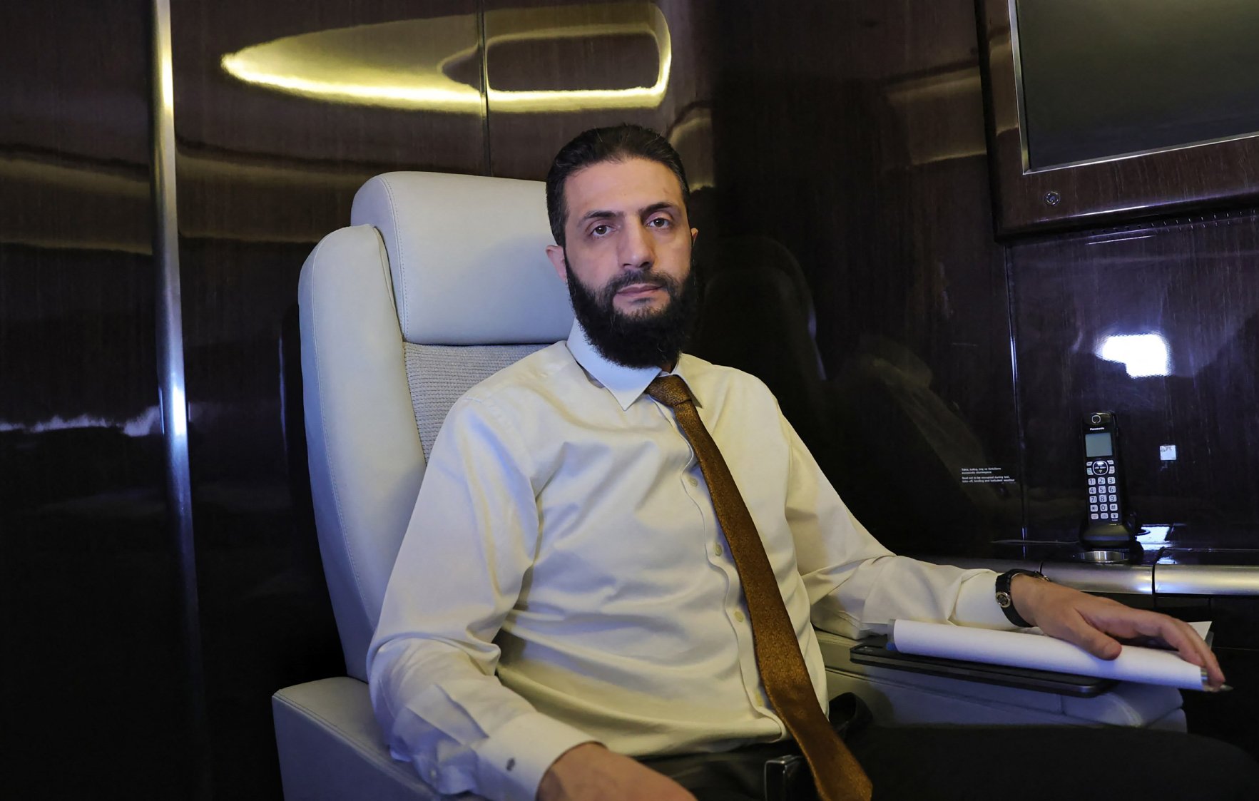 الصورة تُظهر شخصًا جالسًا في مقعد مريح داخل مكان يبدو راقيًا. يرتدي قميصًا فاتحًا ورابطة عنق، ويبدو في وضعية تفكر. خلفه توجد شاشة ومكتب أنيق، مما يوحي بأجواء مهنية.