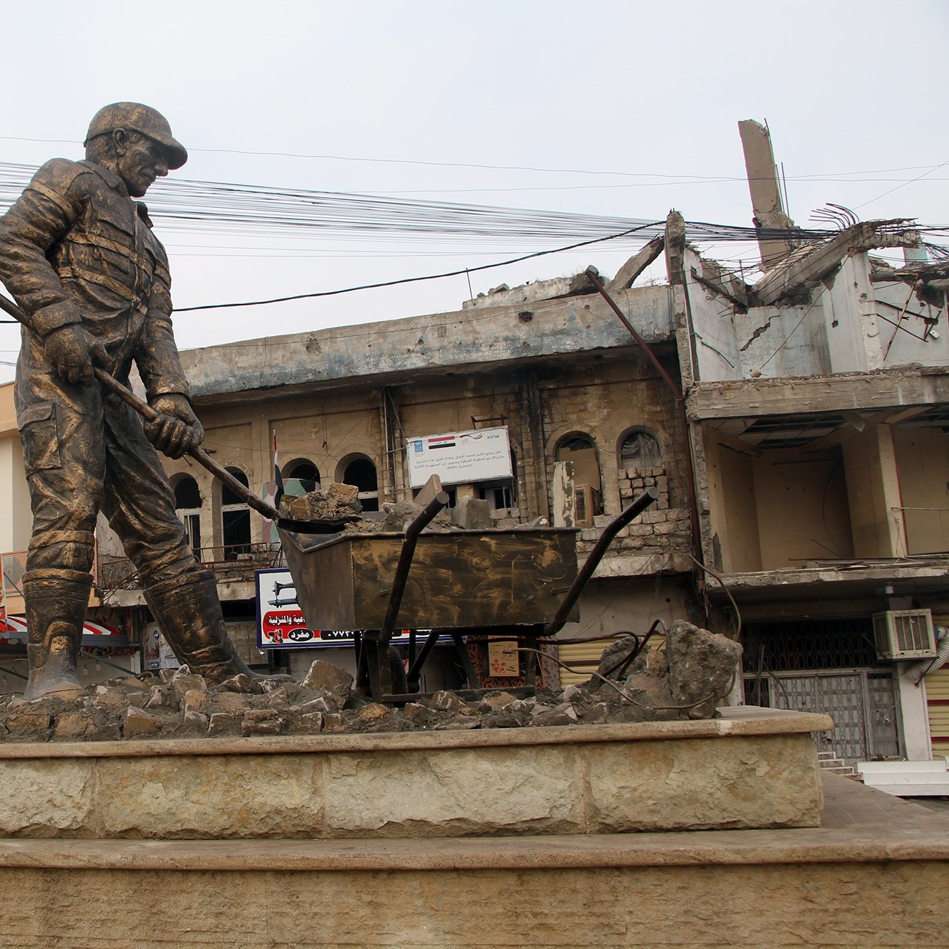 L'image montre une statue monumentale représentant un travailleur en train de creuser, symbolisant probablement la reconstruction ou le labeur dans un contexte difficile. En arrière-plan, on distingue des bâtiments partiellement détruits et des infrastructures urbaines. La scène évoque une atmosphère de résilience et de reconstruction dans une zone touchée par des conflits. La statue est installée sur un socle en pierre, ajoutant à son importance et à son impact visuel.
