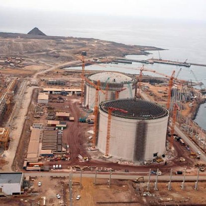 L'image montre un site industriel en construction, probablement une installation de gaz naturel ou une centrale électrique. On peut voir plusieurs grands réservoirs cylindriques, entourés de grues et de matériaux de construction. L'environnement est côtier, avec une vue sur la mer et des montagnes en arrière-plan. Il y a également des routes et des véhicules de construction présents sur le site. Le paysage est principalement désertique et montagneux.