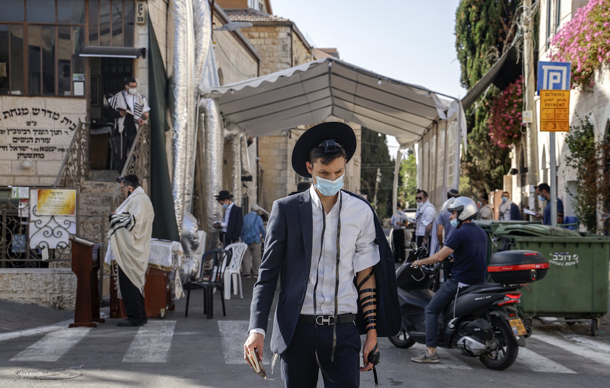L'image montre un homme marchant dans une rue, portant un costume noir et un chapeau. Il a un masque facial et est vêtu d'un tefillin sur son bras. En arrière-plan, on peut voir un endroit semblant être un lieu de prière, avec des gens habillés en vêtements traditionnels. Des panneaux et de la végétation décorent le paysage urbain. La scène évoque une atmosphère communautaire et religieuse.