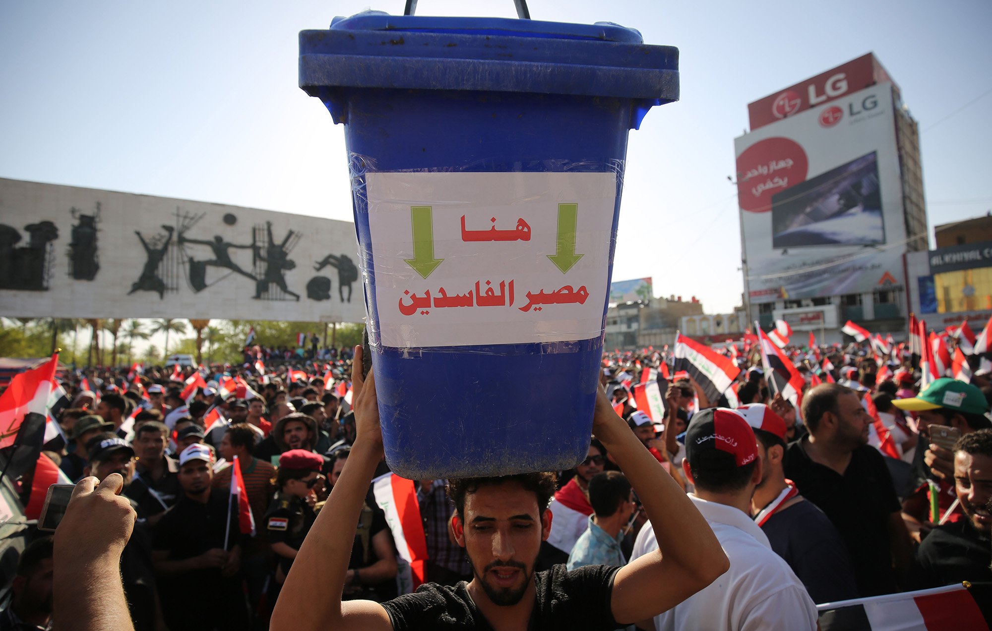L'image montre une grande manifestation avec une foule rassemblée. Au premier plan, un homme porte un grand seau bleu sur sa tête, qui affiche des inscriptions en arabe. Derrière lui, on peut voir de nombreux drapeaux, probablement ceux de l'Égypte, et des affiches graphiques sur un mur. La scène dégage une atmosphère de protestation, avec des gens mobilisés pour une cause.