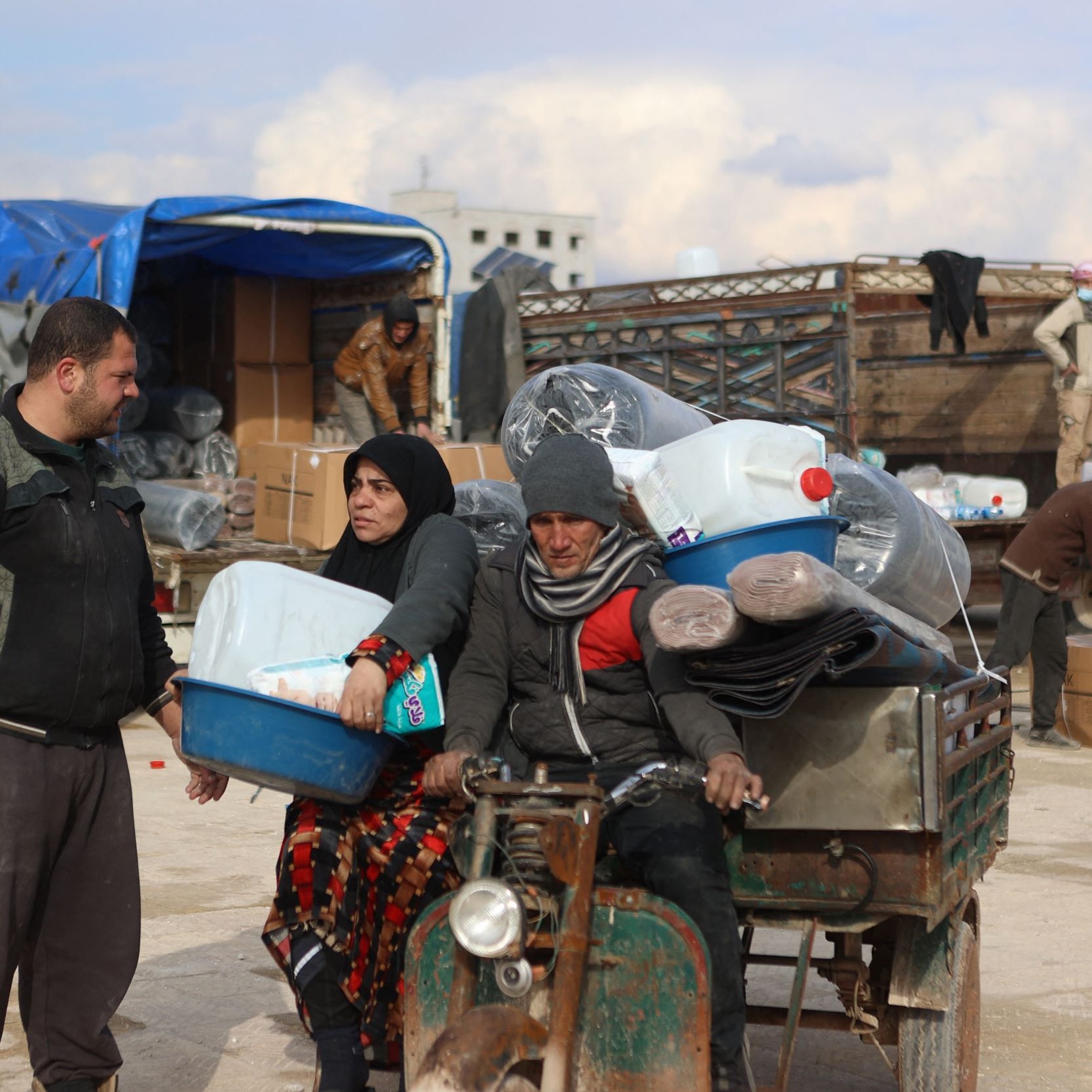 Nell'immagine si vede una scena di un'area di stoccaggio o mercato. Ci sono alcune persone che sembrano occupate a scaricare e trasportare materiali. Un uomo e una donna sono in primo piano: l'uomo indossa abiti scuri e tiene una cassetta, mentre la donna è vestita con un lungo mantello scuro e apparente una certa preoccupazione sul volto. Dietro di loro, ci sono merci come bottiglie di plastica e tappeti, oltre a veicoli e altri uomini che svolgono attività di carico e scarico. L'atmosfera sembra essere quella di un'operazione di supporto umanitario.
