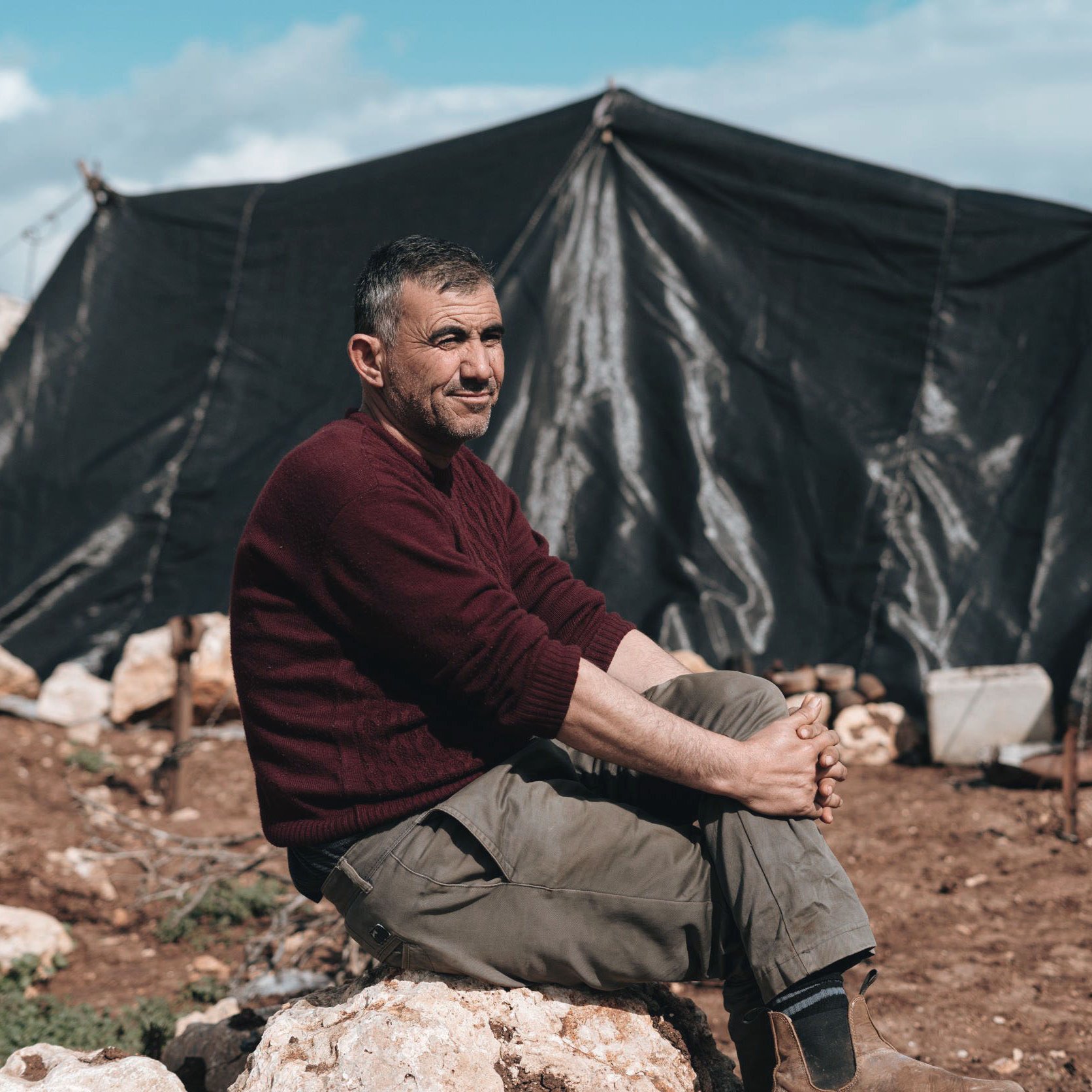 Nell'immagine si vede un uomo seduto su una roccia in un ambiente all'aperto, con un'espressione rilassata. Indossa un maglione scuro e pantaloni chiari. Dietro di lui si nota un grande tendone nero che fa da rifugio, immerso in un paesaggio di terra e rocce. Il cielo è parzialmente nuvoloso, dando un'atmosfera di calma all'ambiente circostante.