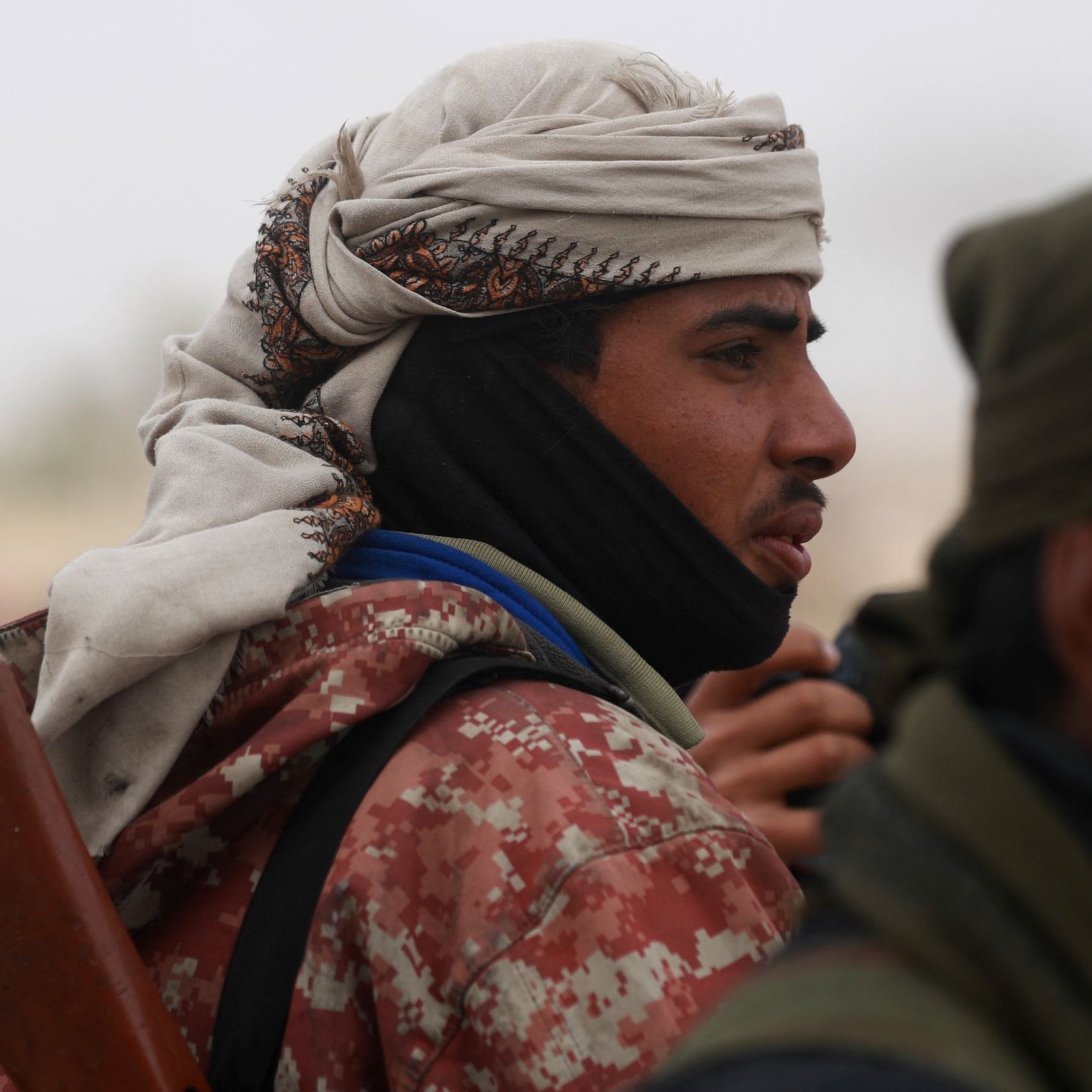 L'image montre un groupe d'individus, dont l'un est au premier plan. Il porte un couvre-chef traditionnel et une écharpe, ce qui lui donne un aspect distinctif. Son expression est sérieuse, et il semble concentré. Il tient également ce qui semble être une arme, indiquant un contexte potentiellement militaire ou de conflit. L'arrière-plan est flou, avec d'autres personnes visibles, suggérant une atmosphère de rassemblement ou d'affrontement.