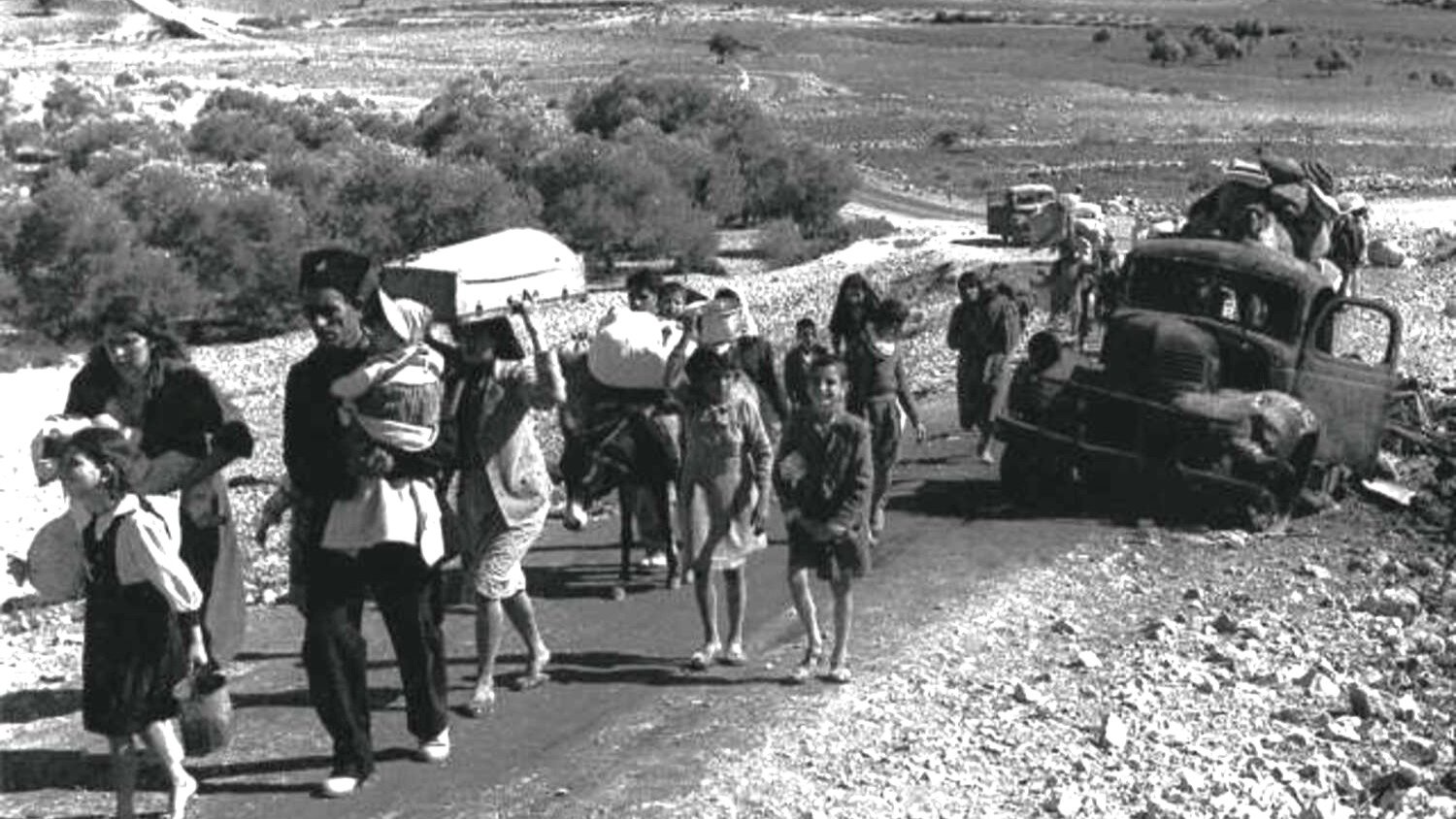 L'image montre un groupe de personnes marchant le long d'une route, probablement en situation de déplacement ou d'exode. On peut voir des adultes portant des enfants et des charges, tandis que des enfants marchent également. À l'arrière-plan, des véhicules abandonnés et un paysage aride s'étendent, suggérant une situation difficile. L'atmosphère semble grave et chargée d'émotion, témoignant des épreuves que traversent ces personnes.