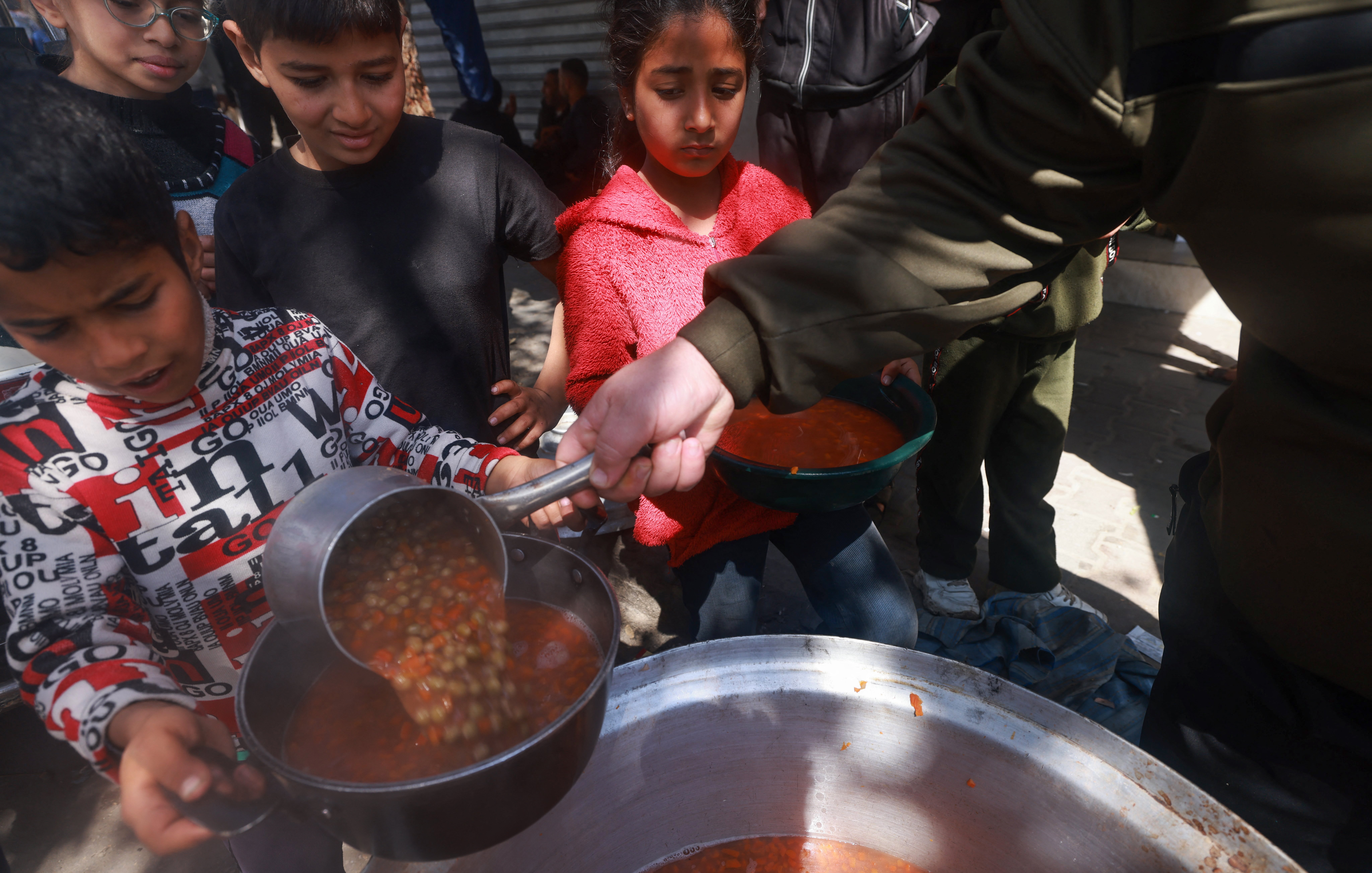 L'immagine mostra un gruppo di bambini in un ambiente all'aperto, mentre qualcuno sta servendo del cibo da una grande pentola. Un bambino, in particolare, sta utilizzando un mestolo per prendere una porzione di una zuppa o stufato, che sembra contenere legumi. Gli altri bambini attendono, con espressioni curiose e attente, alcuni con uno sguardo serio. L'atmosfera trasmette un senso di comunità e condivisione, evidenziando un momento significativo di aiuto e sostegno.