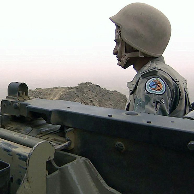 L'image montre un soldat en uniforme, portant un casque, positionné à côté d'une mitrailleuse. Il semble surveiller son environnement avec une expression concentrée. Le paysage derrière lui est brumeux, ce qui donne une atmosphère de tension et de vigilance. Le soldat est en posture d'attente, prêt à agir si nécessaire.