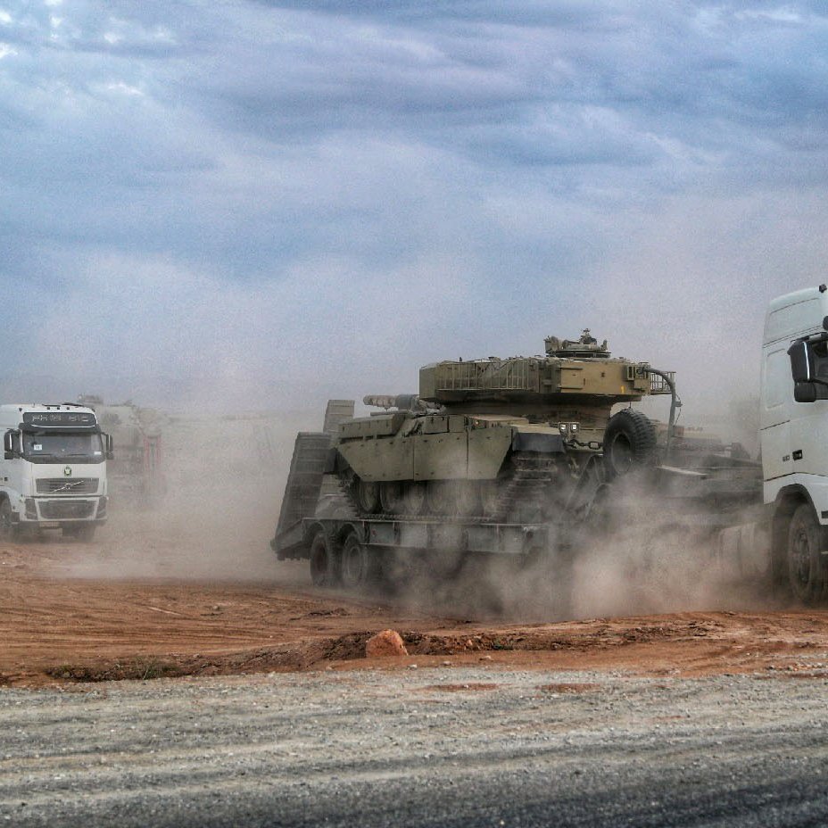 L'image montre une scène de transport militaire, avec plusieurs camions, notamment un portant le logo Volvo, transportant des véhicules blindés. La poussière s'élève autour des véhicules, suggérant un environnement saharien ou aride. Le ciel est nuageux, ajoutant à l'atmosphère de la scène. Les camions avancent sur une route en terre, mettant en avant un aspect logistique lié aux opérations militaires.