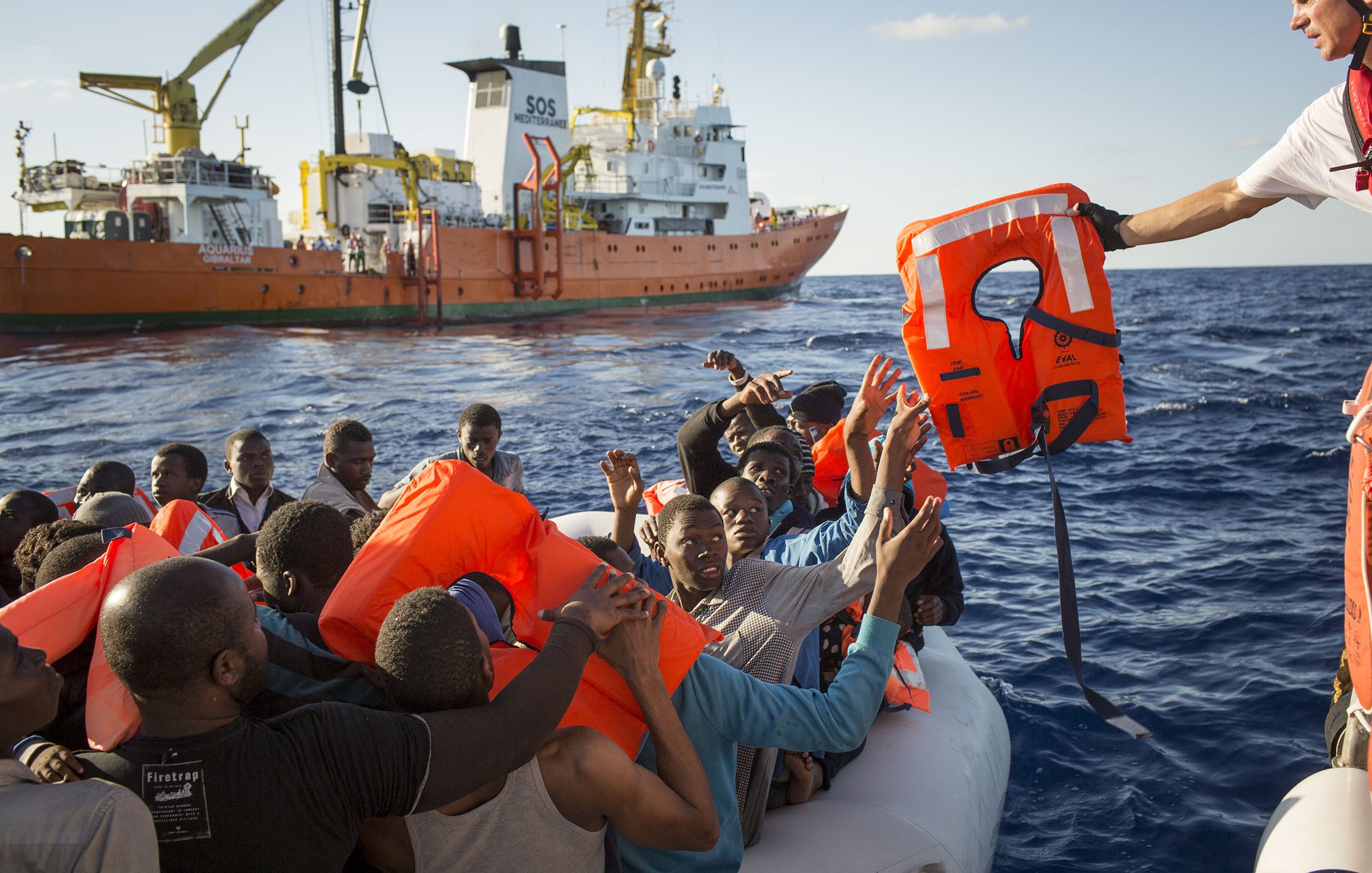 L'image montre une scène en mer où un groupe de personnes se trouve sur un canot pneumatique. Elles semblent en détresse et soulèvent les bras en direction d'un sauveteur qui leur tend une bouée de sauvetage. En arrière-plan, un grand navire de secours est visible, avec des couleurs orange et blanc. L'atmosphère semble tendue, et on peut ressentir l'urgence de la situation. Le ciel est dégagé et l'eau est calme, contrastant avec la gravité du moment.