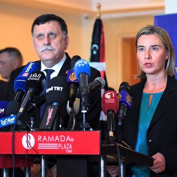 تظهر الصورة مؤتمرًا صحفيًا حيث تقف امرأة أمام مجموعة من الميكروفونات، بينما يقف رجل خلفها. المرأة تبدو وكأنها تتحدث بجدية، بينما خلفها يوجد علم للدولة، بالإضافة إلى لافتة تحمل شعار الاتحاد الأوروبي. الأضواء مركزة على المتحدثين، مع وجود مجموعة من الإعلاميين في الخلفية.