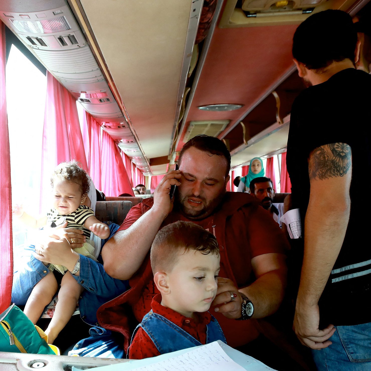 L'image montre un intérieur de bus avec plusieurs personnes. Un homme assis au centre tient un enfant dans ses bras et parle au téléphone. À ses pieds, un autre enfant semble jouer ou être attentif. D'autres passagers sont visibles autour, certains se tenant debout. Les sièges sont occupés, et des rideaux rouges laissent passer une lumière douce à travers les fenêtres. L'atmosphère semble chaleureuse et animée.