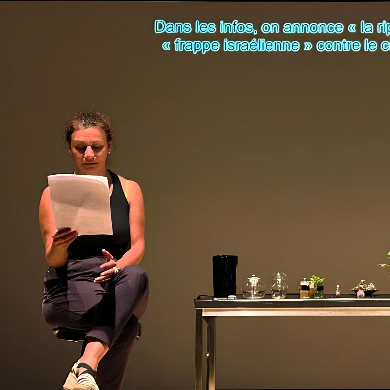 Una donna legge appunti davanti a un tavolo con piante e oggetti, su uno sfondo neutro.