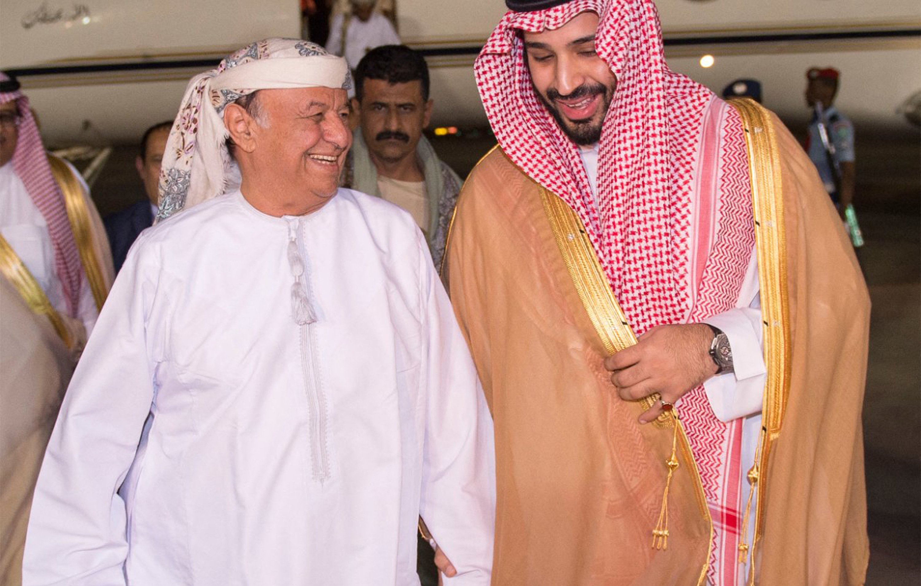 في الصورة، يمكن رؤية رجلين يتحدثان معاً بمرح. يرتدي أحدهما زياء تقليدية عربية مكونة من جبة بيضاء وعقال، بينما يرتدي الآخر الملابس السعودية التقليدية، تشمل العباءة والكوفية. يظهر خلفهم طائرة خاصة، مما يشير إلى وصولهم إلى مكان محدد. تبدو الأجواء مفعمة بالترحاب والود.