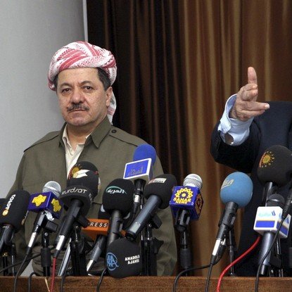 L'image montre deux hommes en conférence de presse, entourés de plusieurs micros et caméras provenant de divers médias. L'homme à gauche porte une tenue traditionnelle kurde avec un foulard, tandis que l'homme à droite, habillé en costume, semble s'adresser à l'audience en pointant du doigt. L'ambiance est formelle, indiquant un moment important de communication ou d'annonce.