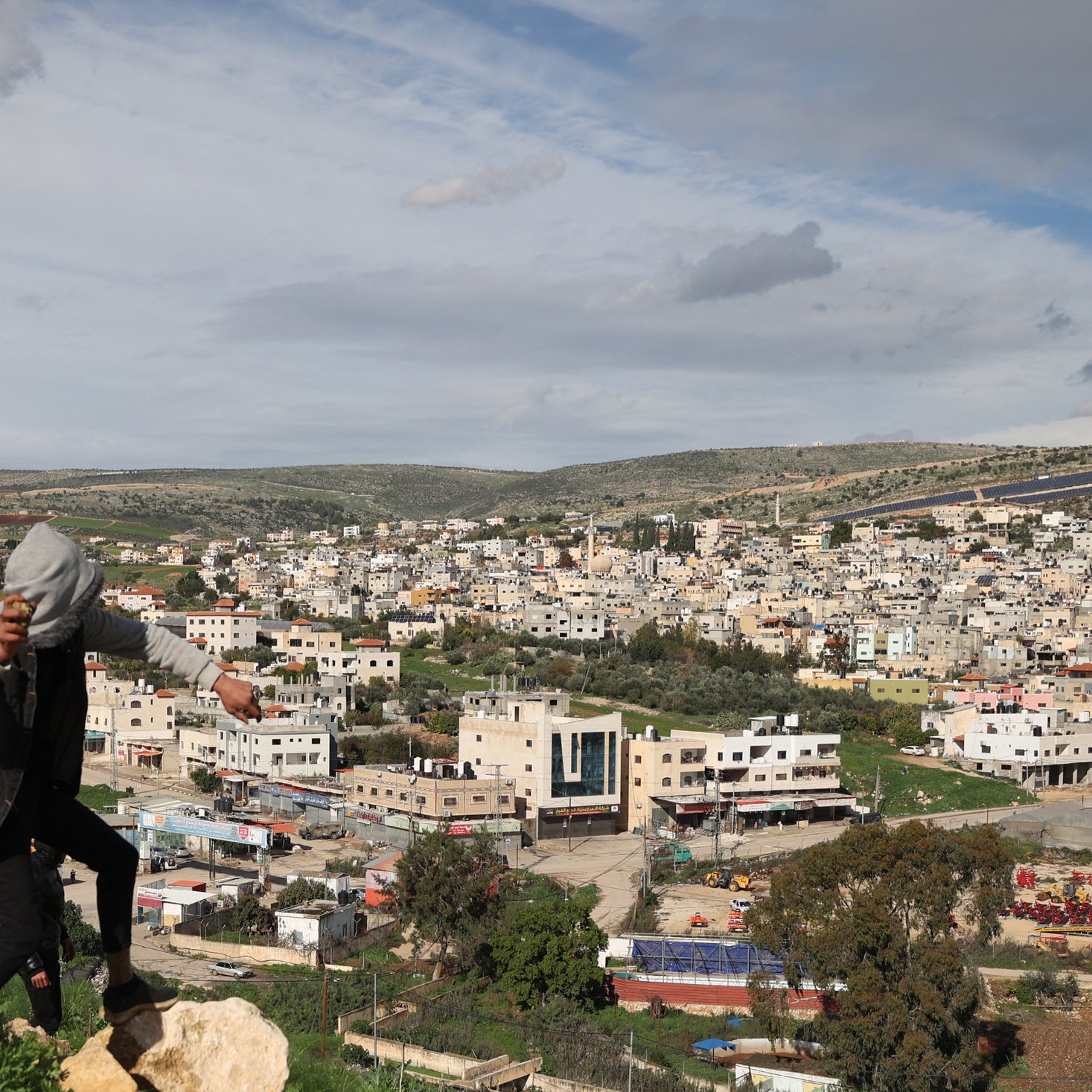 Nell'immagine si vede un giovane che si sta arrampicando su una roccia, con una città sullo sfondo. La città è composta da edifici di diversi piani, molti dei quali di colore chiaro. L'area è circondata da colline verdi e il cielo è parzialmente nuvoloso, suggerendo un clima variabile. Si possono notare anche alcune strutture agricole e aree di terreno aperto in primo piano.