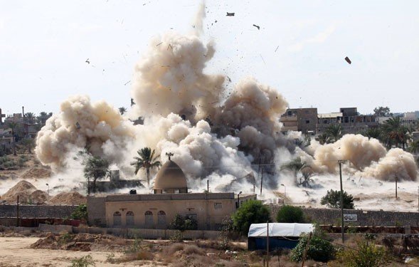 L'image montre une scène d'explosion avec de la fumée et des débris projetés dans les airs. En arrière-plan, on peut apercevoir des bâtiments, des palmiers et un terrain poussiéreux. L'explosion suggère un événement dramatique ou un conflit, et l'environnement semble rural ou semi-urbain. Des signes de destruction sont visibles, soulignant l'impact de l'explosion sur la zone environnante.