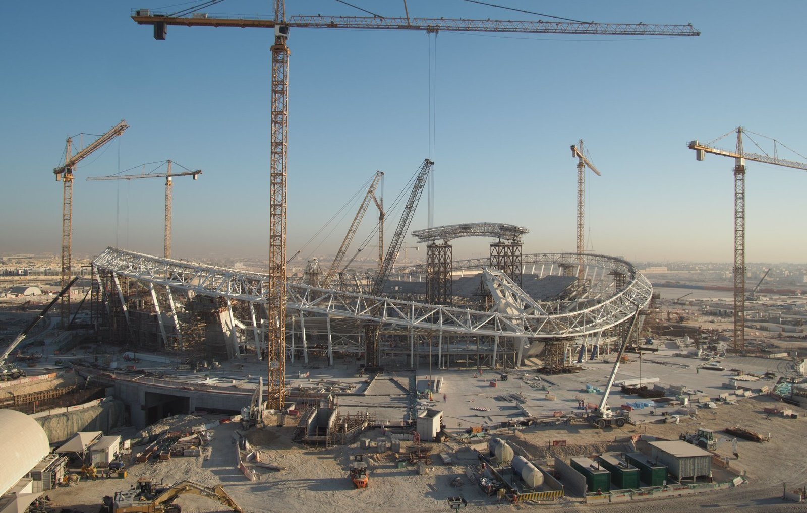 L'image montre un grand chantier de construction d'un stade, avec plusieurs grues en fonctionnement. Le stade a une structure moderne et architecturale, avec des éléments en acier visibles. En arrière-plan, on peut voir un ciel clair, suggérant qu'il fait jour. Le site est entouré de divers matériaux de construction et d'équipements, ce qui indique que le projet est encore en cours.