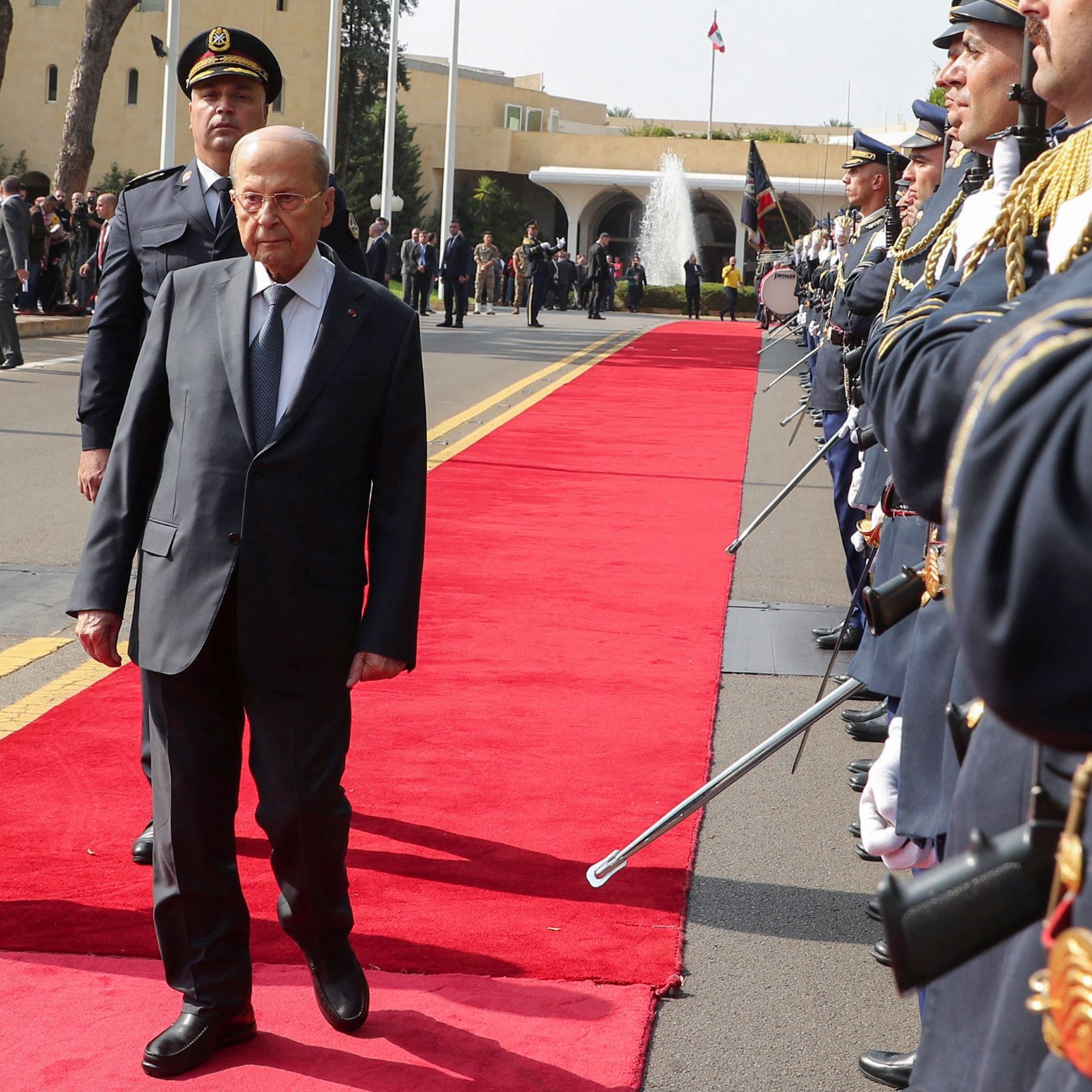 L'image montre une cérémonie officielle. Un homme en costume, probablement une personnalité politique, marche sur un tapis rouge au centre de la scène. De chaque côté, des membres d'une garde d'honneur en uniforme militaire sont alignés, tenant des armes. En arrière-plan, on aperçoit des bâtiments officiels et des drapeaux. L'atmosphère semble solennelle et formelle, suggérant une réception ou un événement diplomatique.