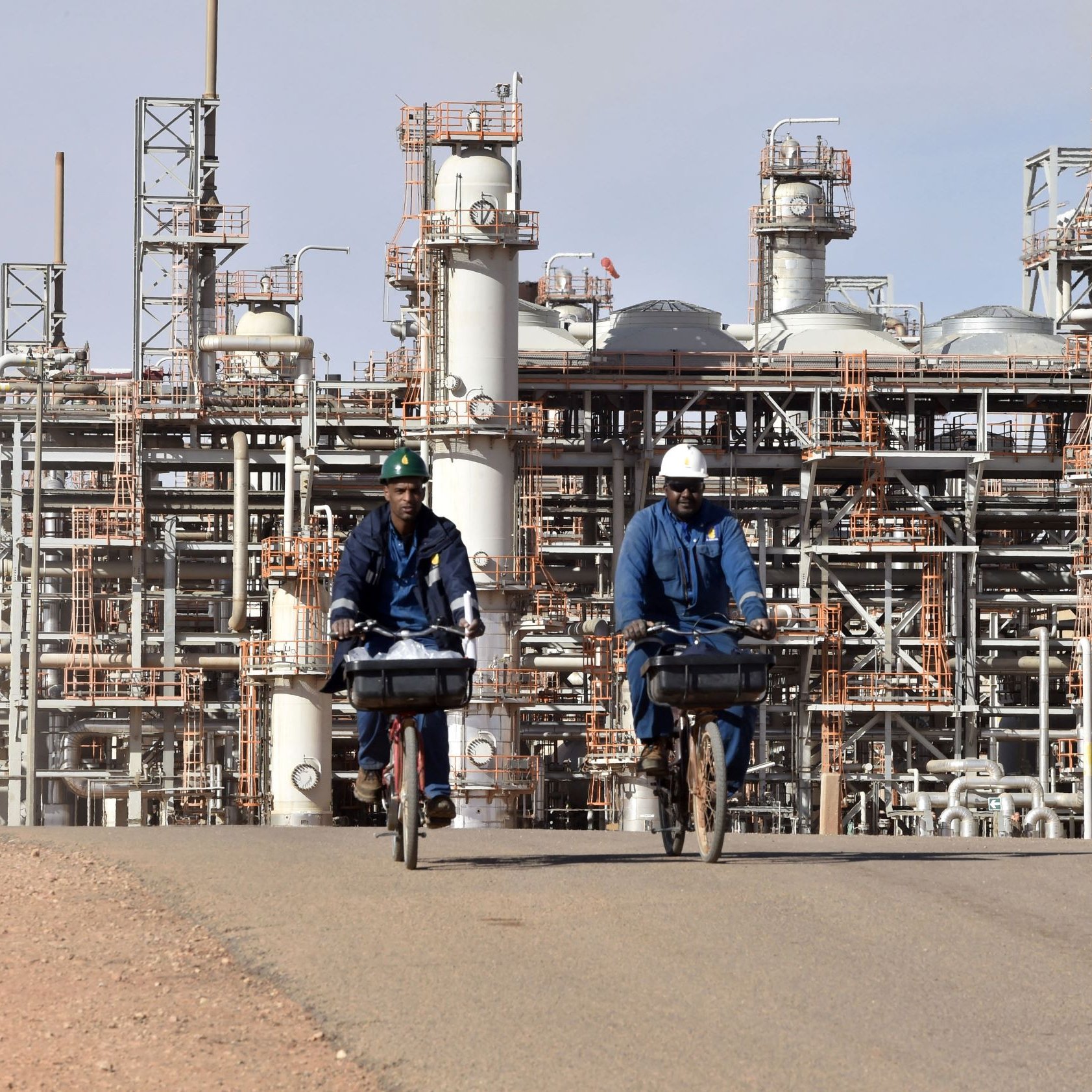 تظهر الصورة مشهدًا من منطقة صناعية، حيث يقوم شخصان يرتديان زي العمل و الخوذة بركوب دراجتين على طريق داخل المصنع. في خلفية الصورة، توجد معدات صناعية كبيرة أنابيب وأبراج مخصصة لعمليات التصنيع أو التكرير. السماء تبدو صافية والجو العام يوحي بنشاطات صناعية قوية.