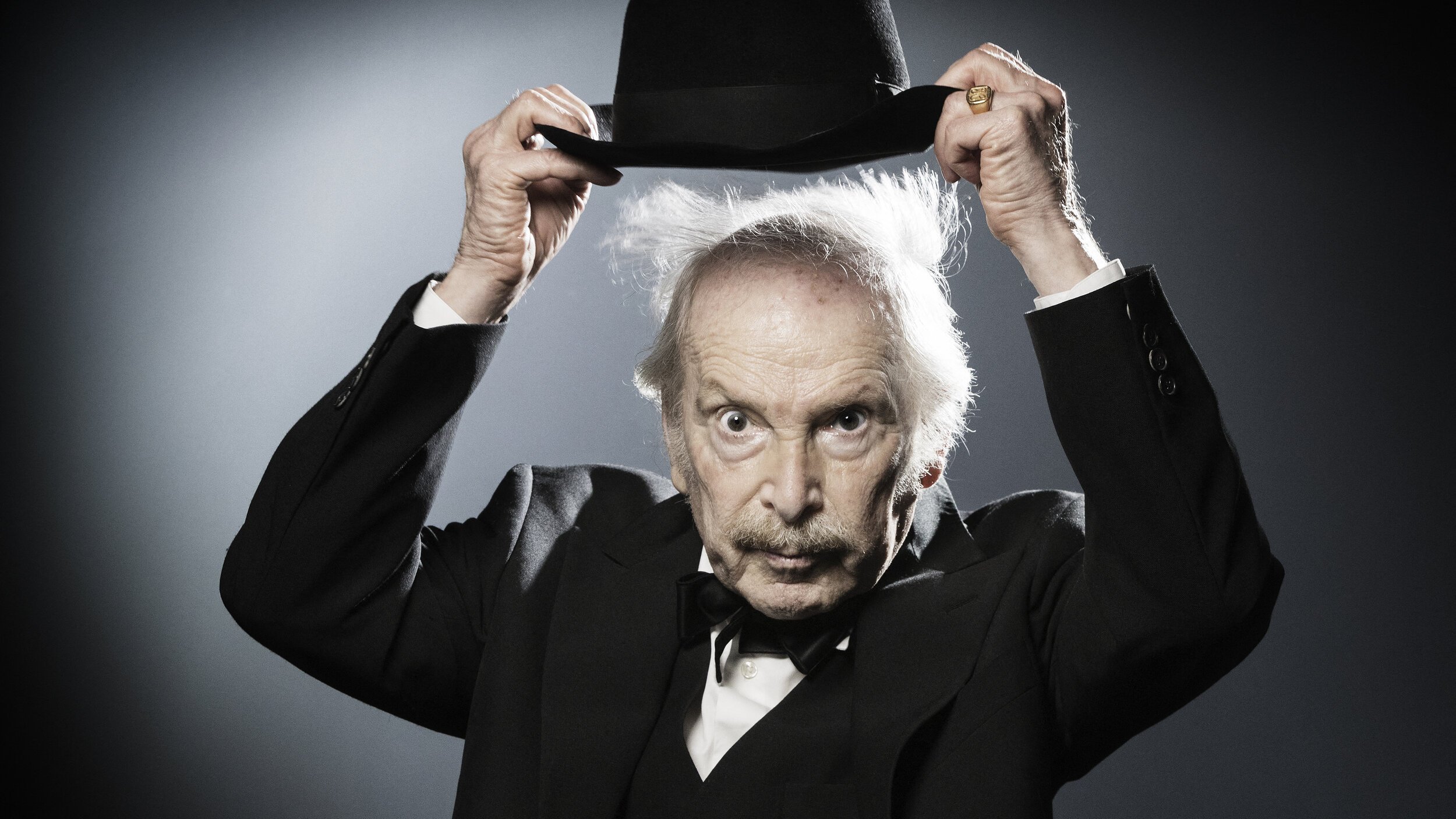 L'image montre un homme âgé portant un costume noir avec un nœud papillon. Il tient un chapeau melon avec une main, et son autre main semble légèrement levée. Ses cheveux sont ébouriffés, ce qui donne un aspect excentrique à son expression. L'arrière-plan est sobre, avec une lumière qui crée un effet dramatique, mettant en valeur son visage et son chapeau.