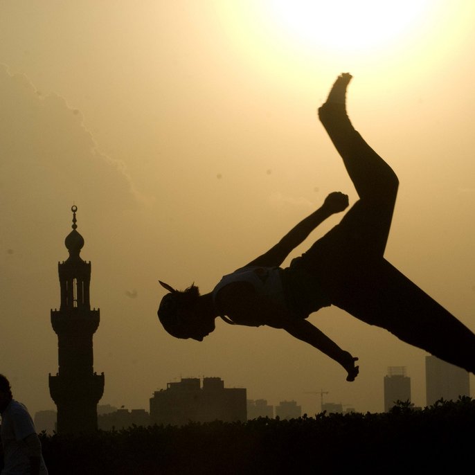 L'image montre une silhouette d'une personne effectuant un saut ou un mouvement acrobatique contre un fond de soleil couchant. À l'arrière-plan, on aperçoit une mosquée avec un minaret, ainsi que des bâtiments de la ville. L'éclairage crée un contraste fort entre la silhouette et le ciel lumineux, ajoutant une atmosphère dynamique et artistique à la scène.