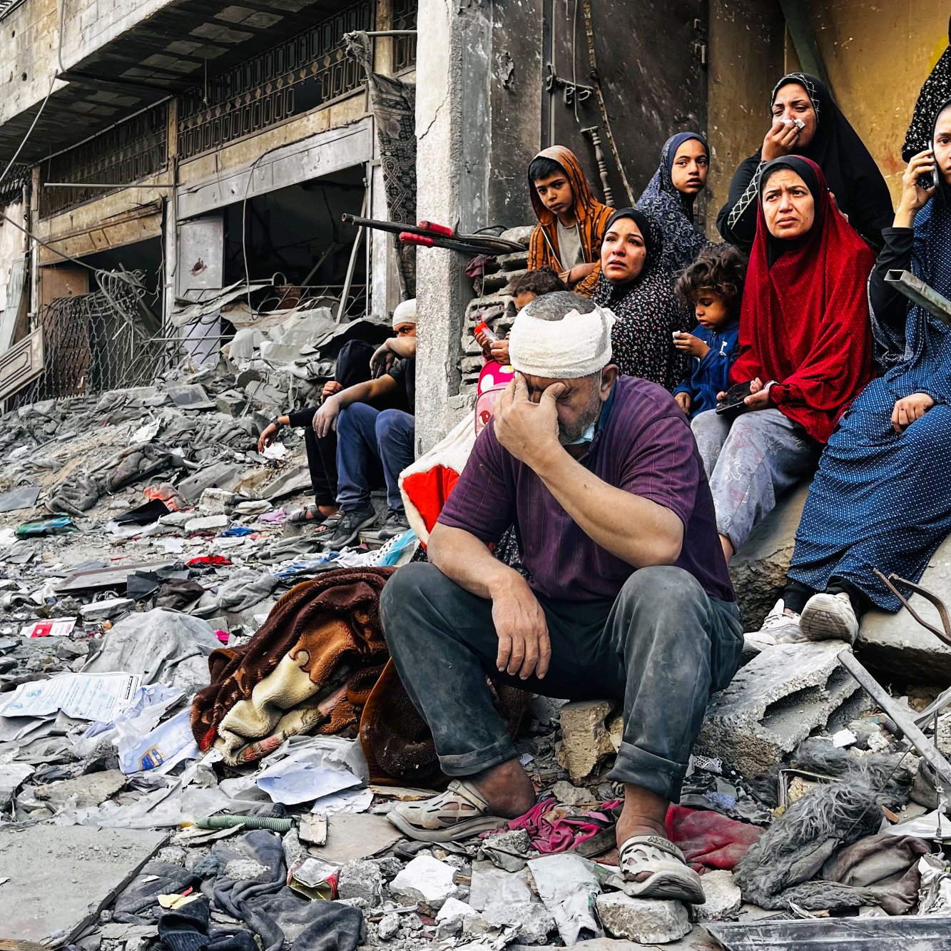 L'immagine mostra una scena devastante in un contesto di guerra o di crisi. Al centro, un uomo con una benda in testa si siede su un cumulo di macerie, con un'espressione di sofferenza e preoccupazione. Attorno a lui, un gruppo di donne sono sedute, vestite con abiti scuri e coprenti, che osservano la scena con sguardi tristi e rassegnati. Un bambino è visibile sulla sinistra, anche lui seduto tra i detriti. Il paesaggio è caratterizzato da edifici distrutti e spazzatura sparsa, evidenziando la desolazione e la disperazione della situazione.