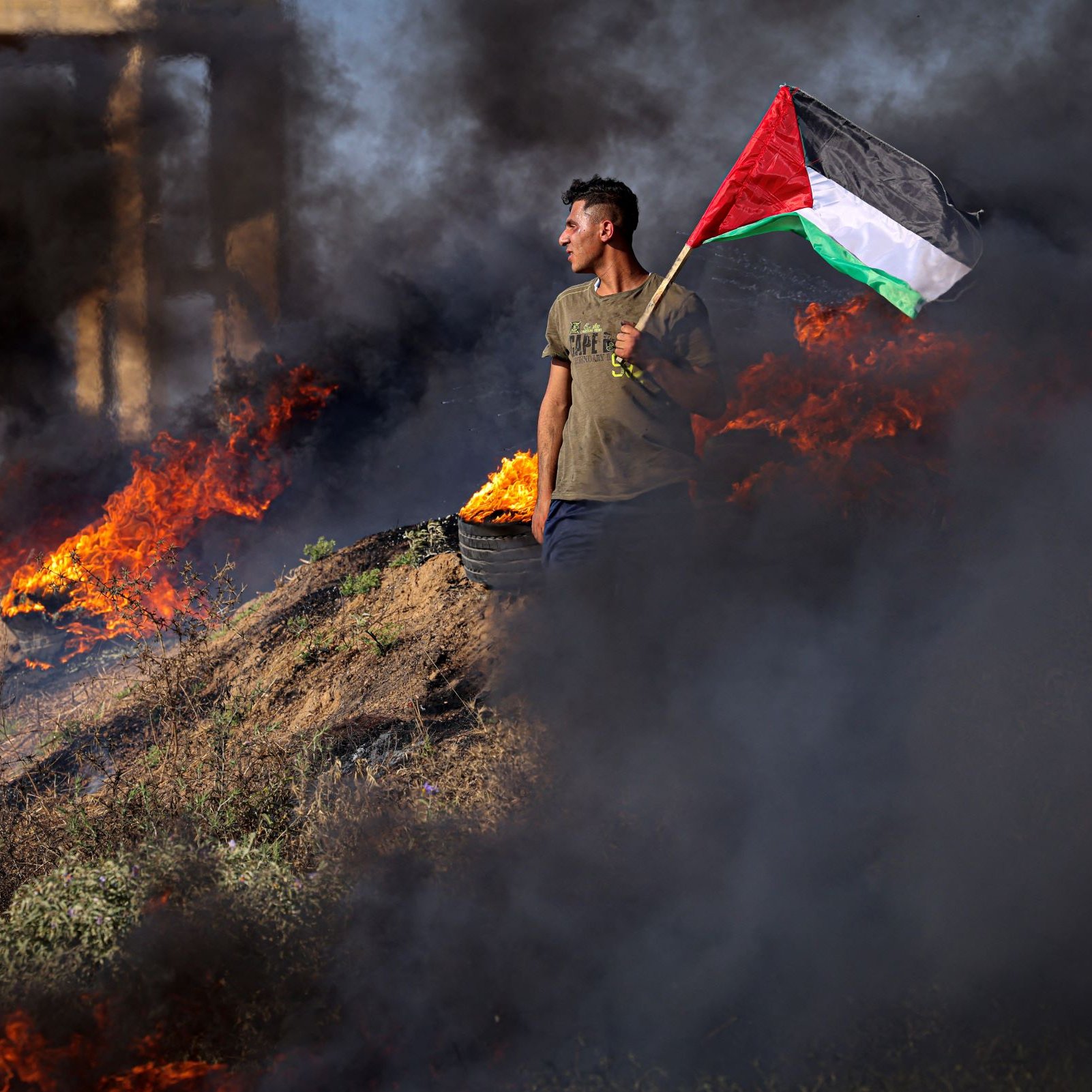 The image depicts a person standing amidst a scene of smoke and flames, holding a Palestinian flag. The individual appears to be in a protest or demonstration setting, surrounded by burning material, which creates a dramatic and intense atmosphere. The contrasting colors of the flag—red, green, black, and white—stand out against the dark smoke and fiery backdrop. The overall mood of the image conveys a sense of defiance and political expression.