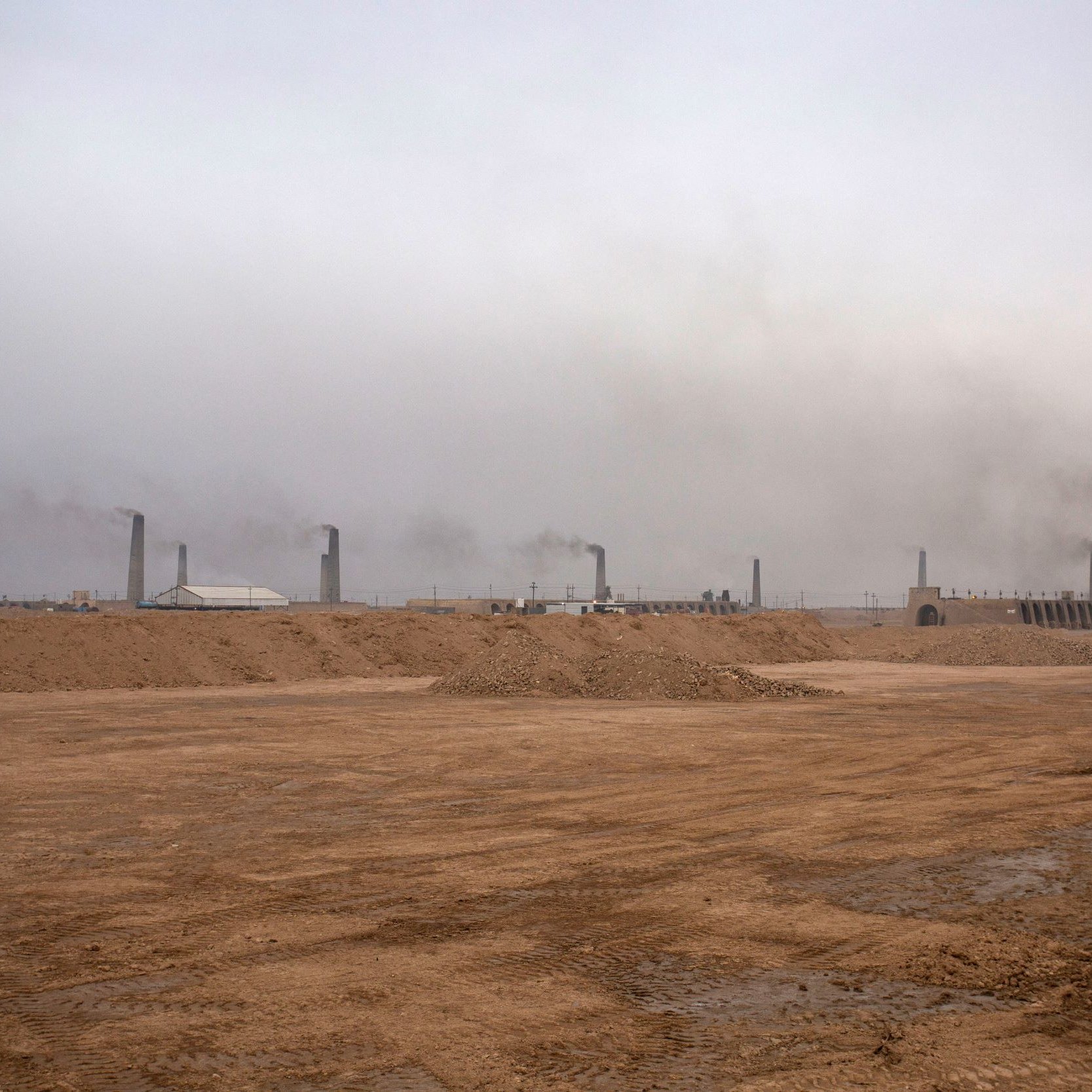 تظهر الصورة منظرًا لموقع صناعي في منطقة صحراوية، حيث يهيمن على المشهد ضباب كثيف أو سحب من الدخان تتصاعد من مداخن المنشآت. الأرض تظهر جافة وغير مستوية، مع كميات من الرمل والتربة. الأجواء تبدو رمادية وموحشة، مما يعكس تأثير النشاط الصناعي على البيئة.
