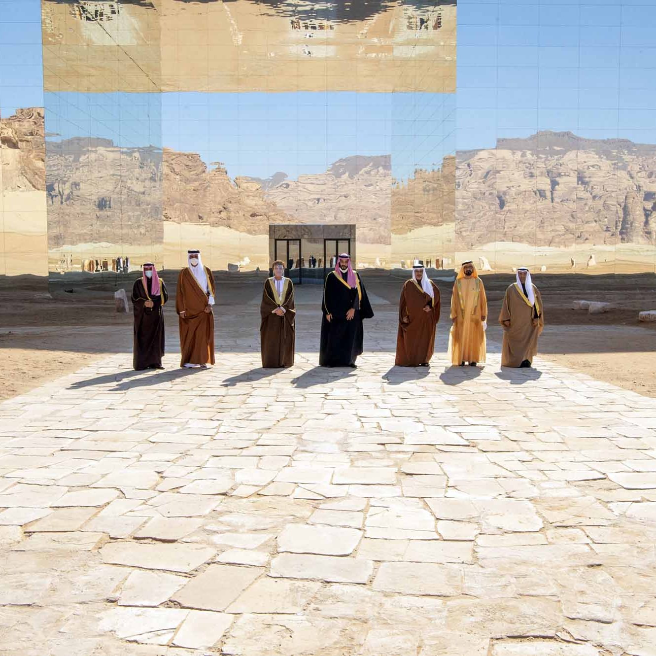 L'image montre un groupe d'individus en vêtements traditionnels, se tenant sur un chemin en pierre au milieu d'un paysage désertique. En arrière-plan, une grande structure réfléchissante fait un miroir du paysage environnant, créant un effet visuel intéressant. Les montagnes et le ciel sont également visibles, ajoutant une dimension naturelle à la scène. L'atmosphère semble solennelle et digne.