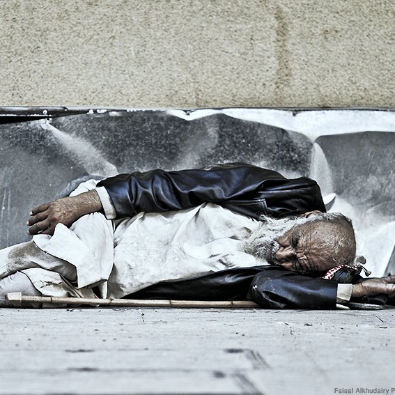 تظهر في الصورة شخصية مسنّة مستلقية على الأرض. يبدو أنه يعاني من الفقر، حيث يرتدي ملابس بسيطة ومتسخة. الوضع الذي هو فيه يعكس مشاعر العزلة والمعاناة. الخلفية تظهر جداراً خشناً، مما يضيف إلى إحساس القسوة في المشهد. تعبر الصورة عن واقع صعب وحياة قاسية يعاني منها البعض في المجتمع.