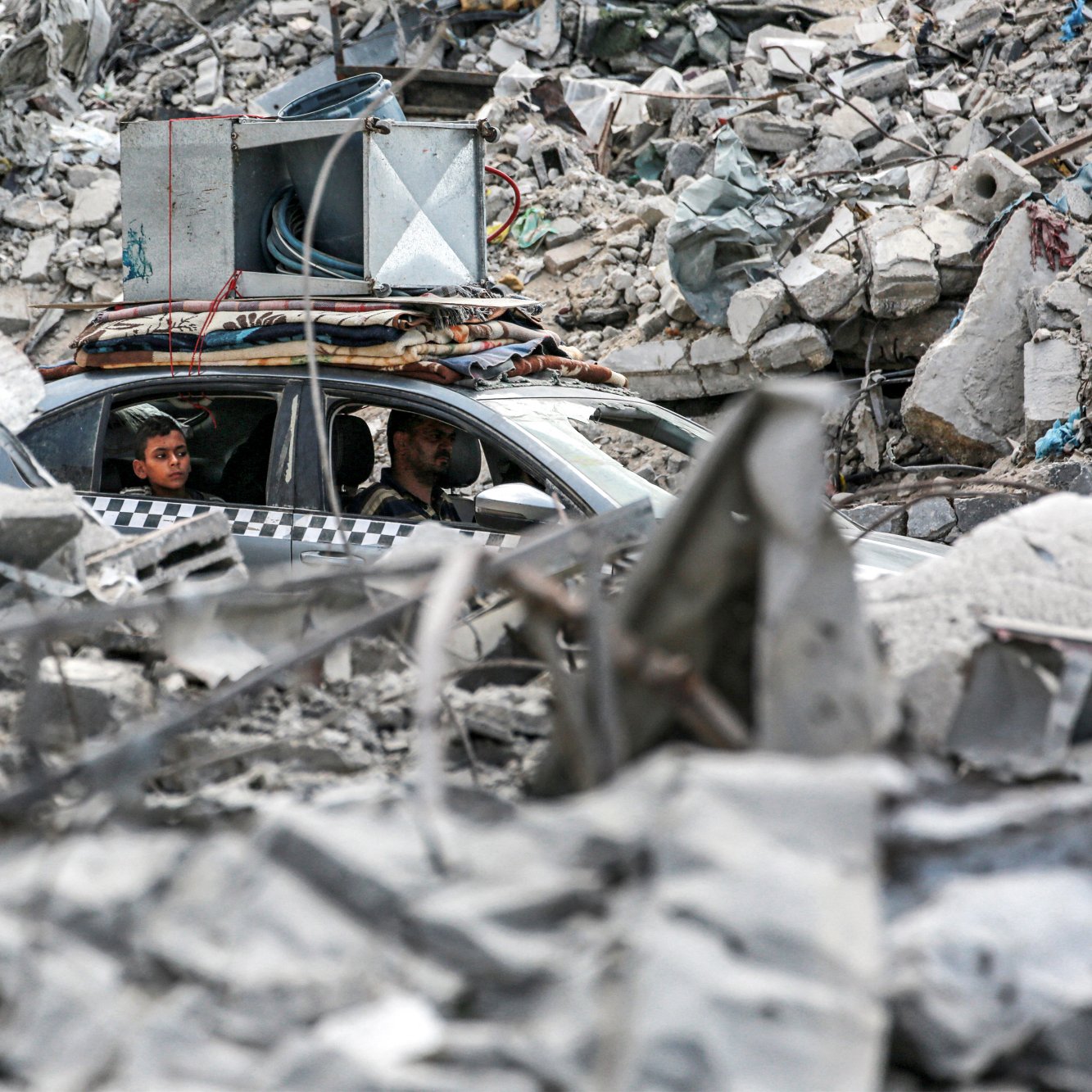 در تصویر، دو مرد درون یک خودرو در میان خرابه‌ها نشسته‌اند. اطراف آن‌ها پر از آوار و بقایای سازه‌های مخروبه است. برخی از أجزاء ساختمانی به وضوح دیده می‌شوند و وضعیتی به شدت آسیب‌دیده را نشان می‌دهند. در بالای خودرو، تجهیزات صوتی قرار گرفته است. فضای کلی تصویر حسی از ویرانی و بحران را منتقل می‌کند.