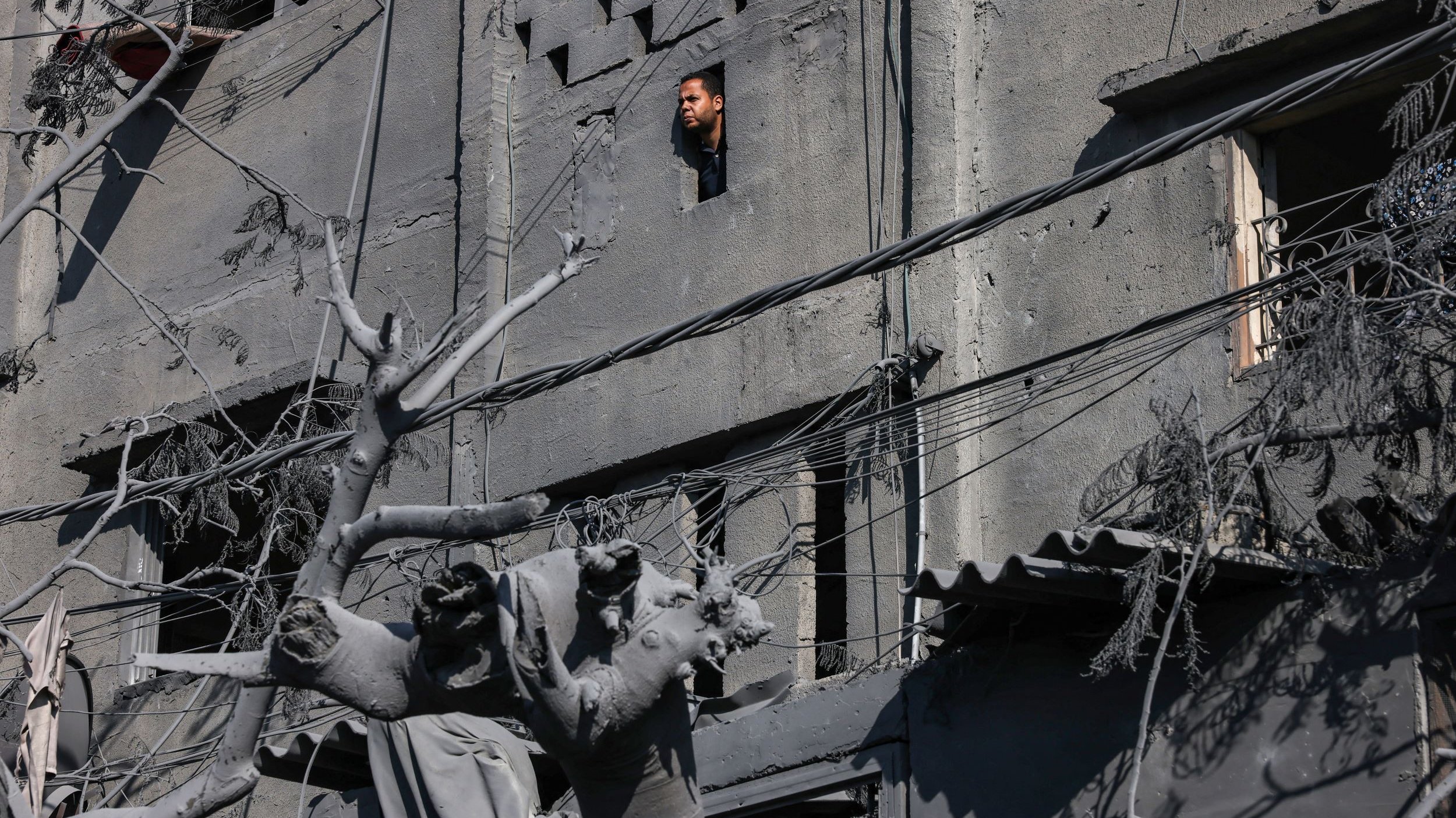 L'image montre un bâtiment en ruines avec des murs de béton gris, où l'on aperçoit une fenêtre. À travers cette fenêtre, une personne regarde vers l'extérieur. Au premier plan, on peut voir des débris et une statue partiellement détruite, entourée de fils électriques et de restes de végétation, ce qui donne une impression de désolation. L'environnement semble touché par un événement tragique, avec une atmosphère sombre et sombre.