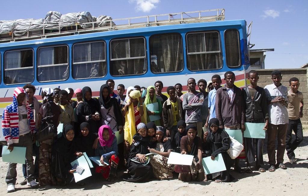 L'image montre un groupe de jeunes adultes et d'adolescents rassemblés devant un bus coloré. Ils semblent être en train de poser pour la photo, tenant tous des documents ou des fichiers. Les personnes portent des vêtements variés, incluant des hijabs et des tenues traditionnelles. L'environnement semble être en plein air, avec un ciel clair et quelques structures visibles en arrière-plan. L'atmosphère paraît conviviale et joyeuse.