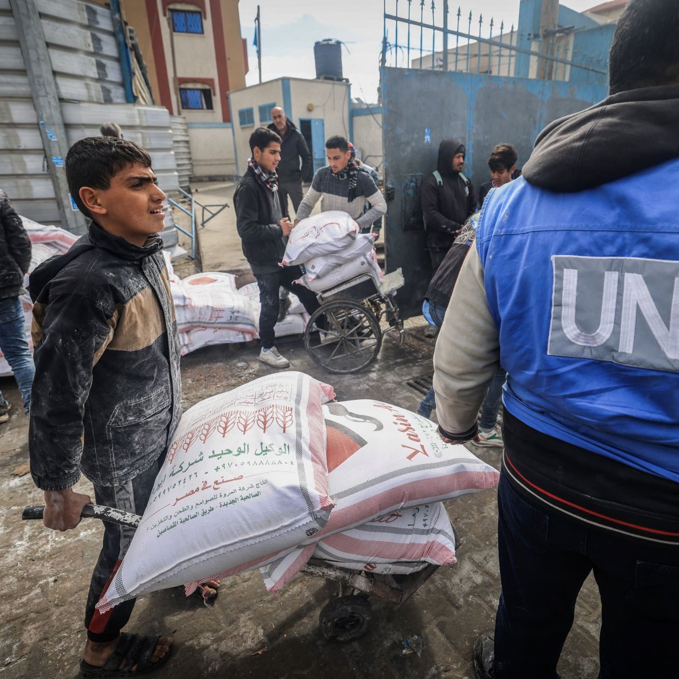 L'immagine mostra una scena di aiuto umanitario. Alcuni ragazzi e adulti sono impegnati nel trasporto di sacchi, probabilmente di cibo o materiali di prima necessità, all'interno di una zona urbana. Un ragazzo sta spingendo un carrello con sacchi, mentre altre persone, alcuni dei quali indossano giubbotti con il logo delle Nazioni Unite, sono visibili in background e stanno trasportando ulteriori carichi. La scena comunica un senso di collaborazione e supporto nella distribuzione di aiuti umanitari.
